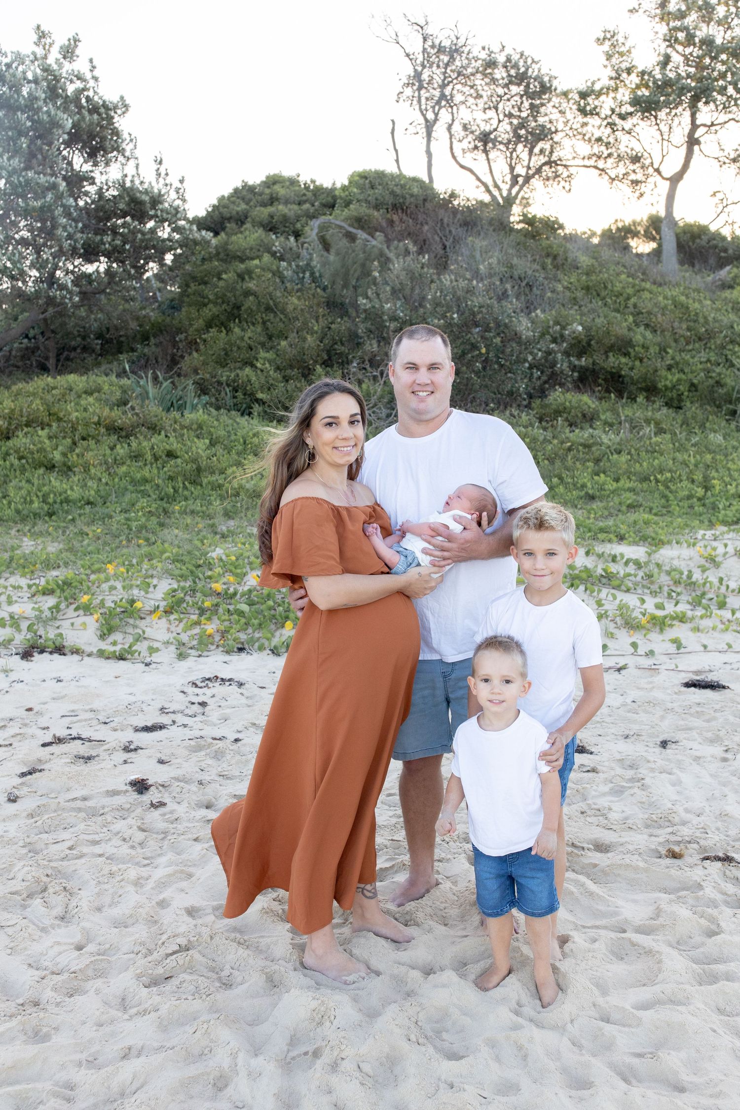 Family of five posing on sandy beach at sunset, father holding newborn, two young boys in white shirts. Coffs Coast photographer.