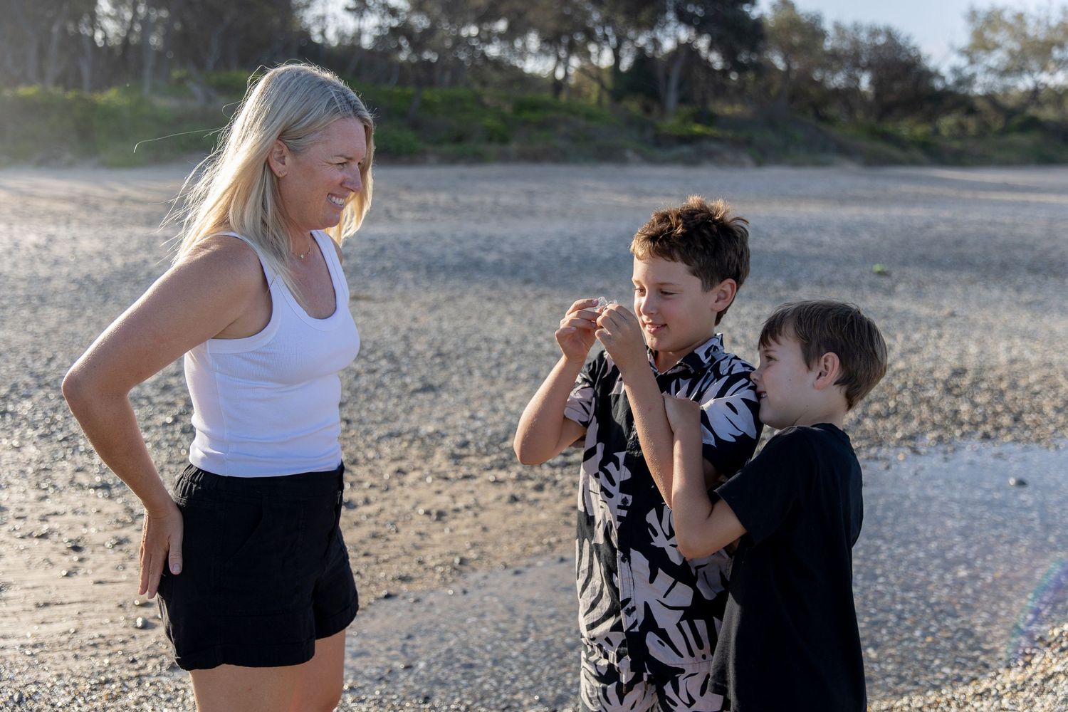 Woman and two boys examining something on a sandy beach during golden hour.