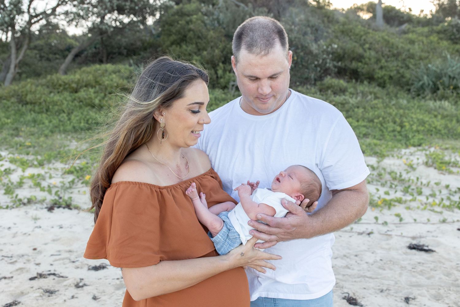 A couple stands on a sandy beach holding their newborn baby, with lush green bushes in the background.