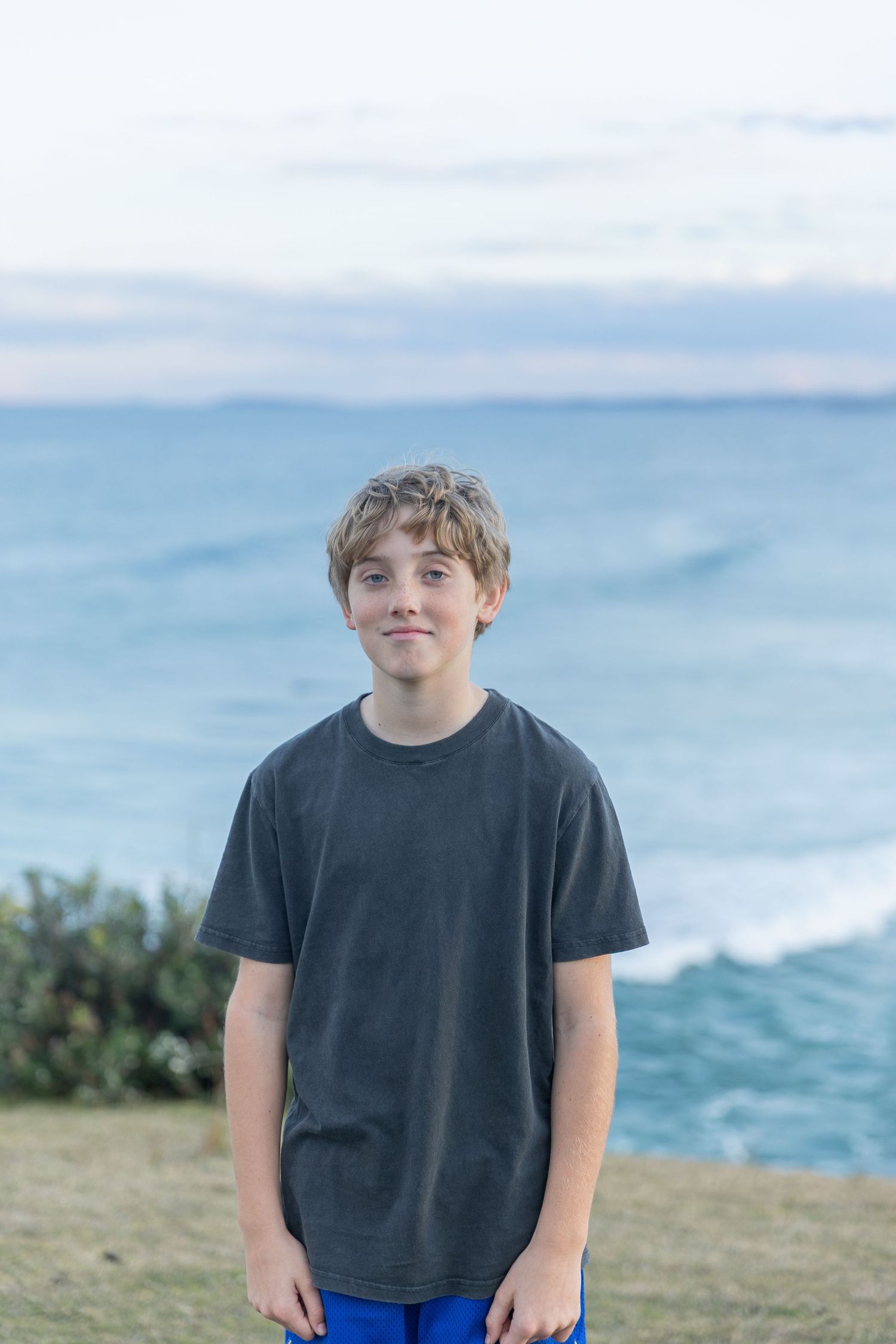 Young person in casual attire standing on coastal hillside with blue ocean background.