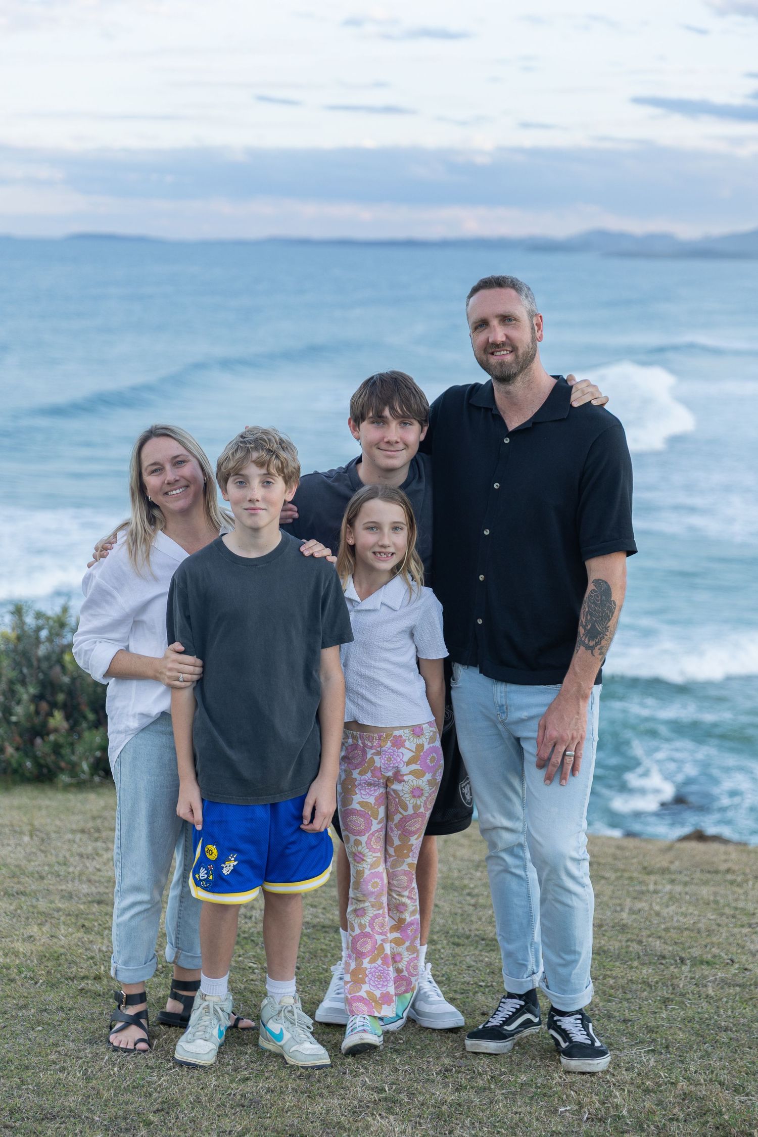 A family group stands together on a grassy coastal cliff overlooking the ocean at sunset.