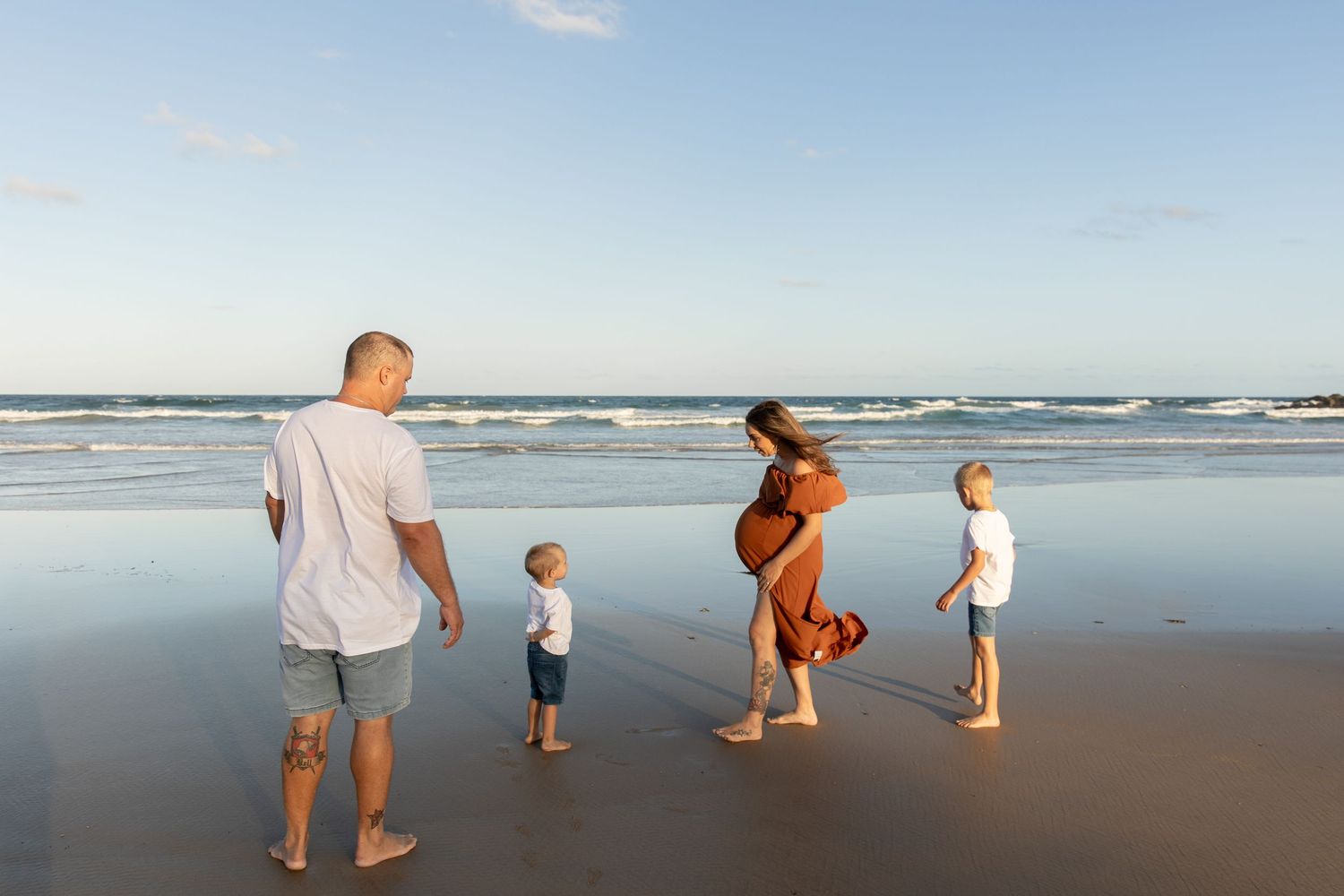 Family of four walking barefoot on a sandy beach at sunset, with a pregnant mother in a rust dress.
