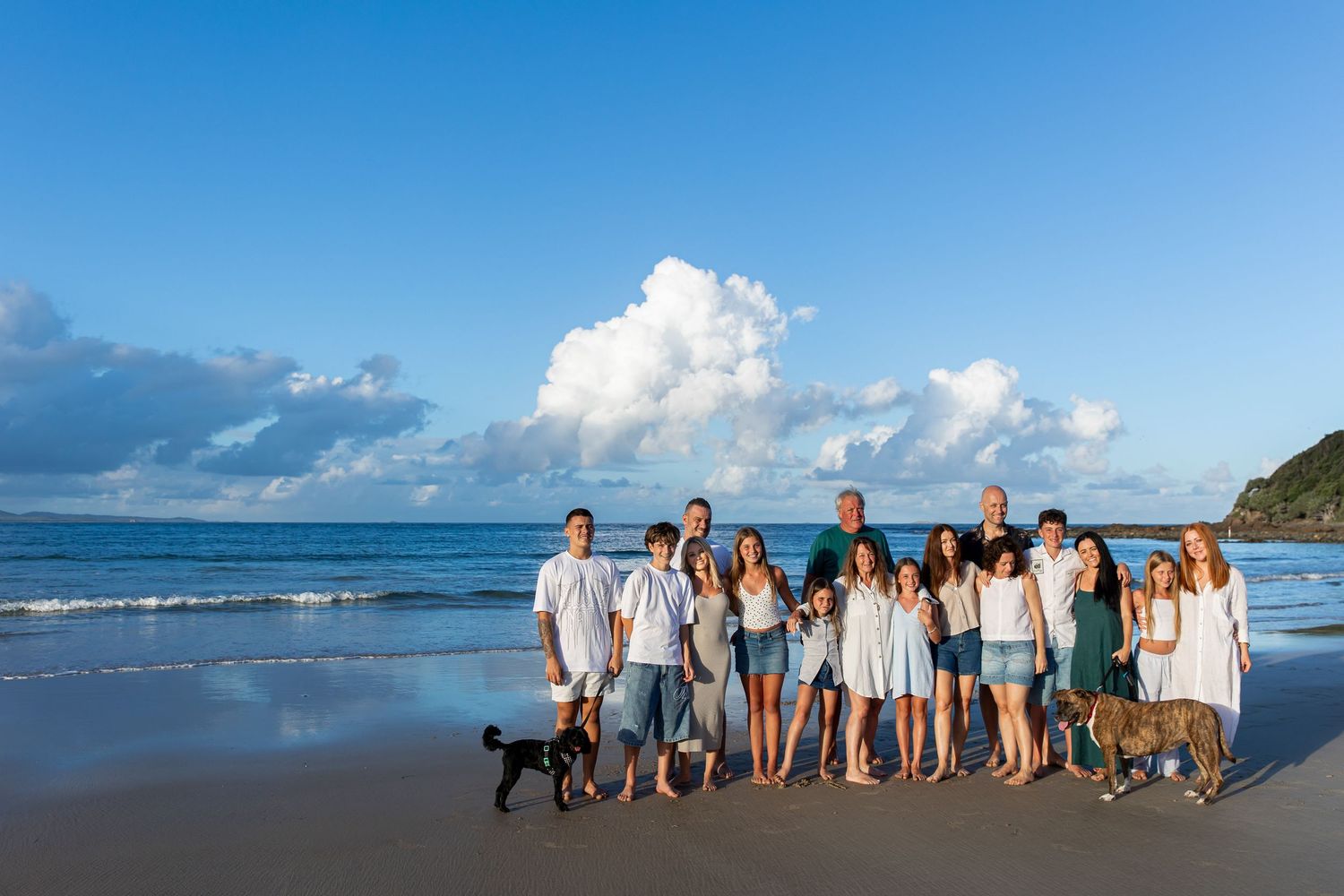Large family group portrait on a tropical beach at golden hour with two dogs, blue sky and white clouds.