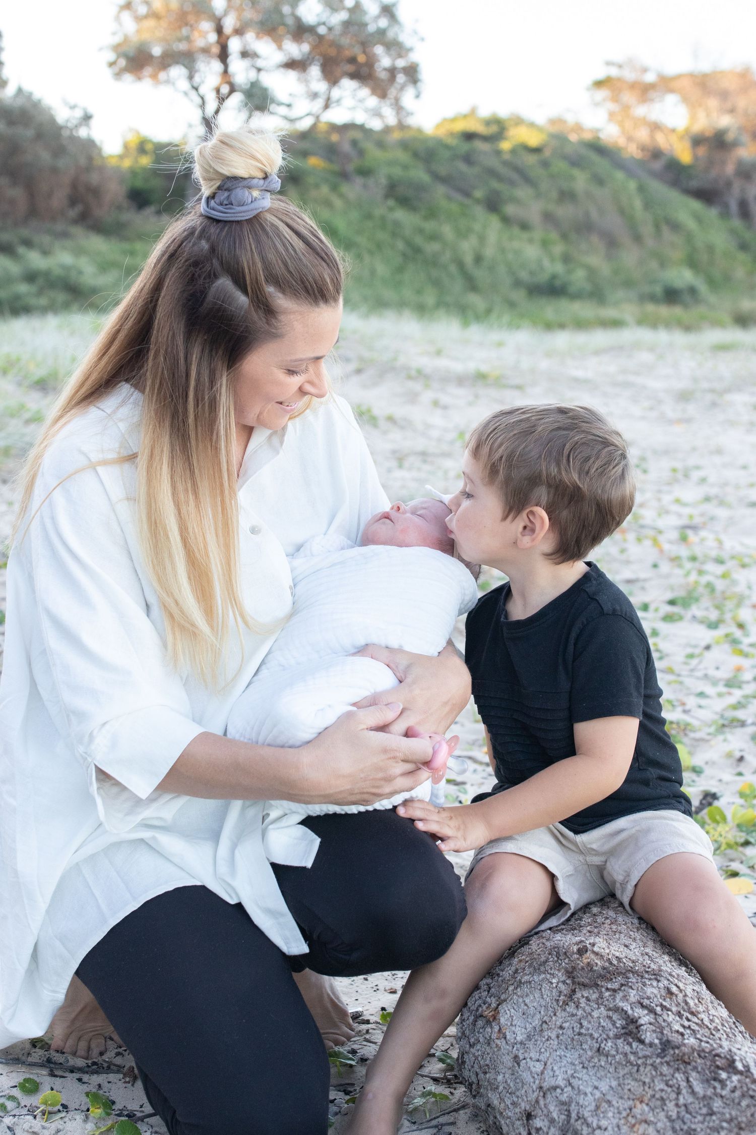 Mother holds newborn baby while young boy leans in to give a gentle kiss on a sandy beach. Coffs Coast photographer.