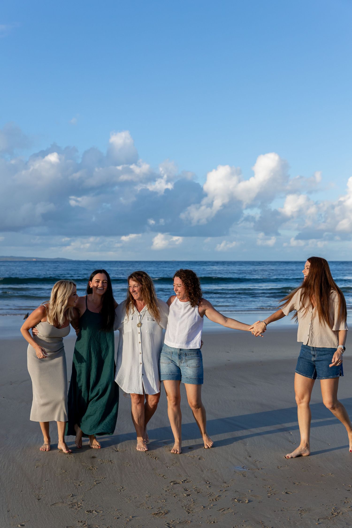 Five women laughing and holding hands on a sandy beach with blue ocean waves and cloudy sky backdrop.