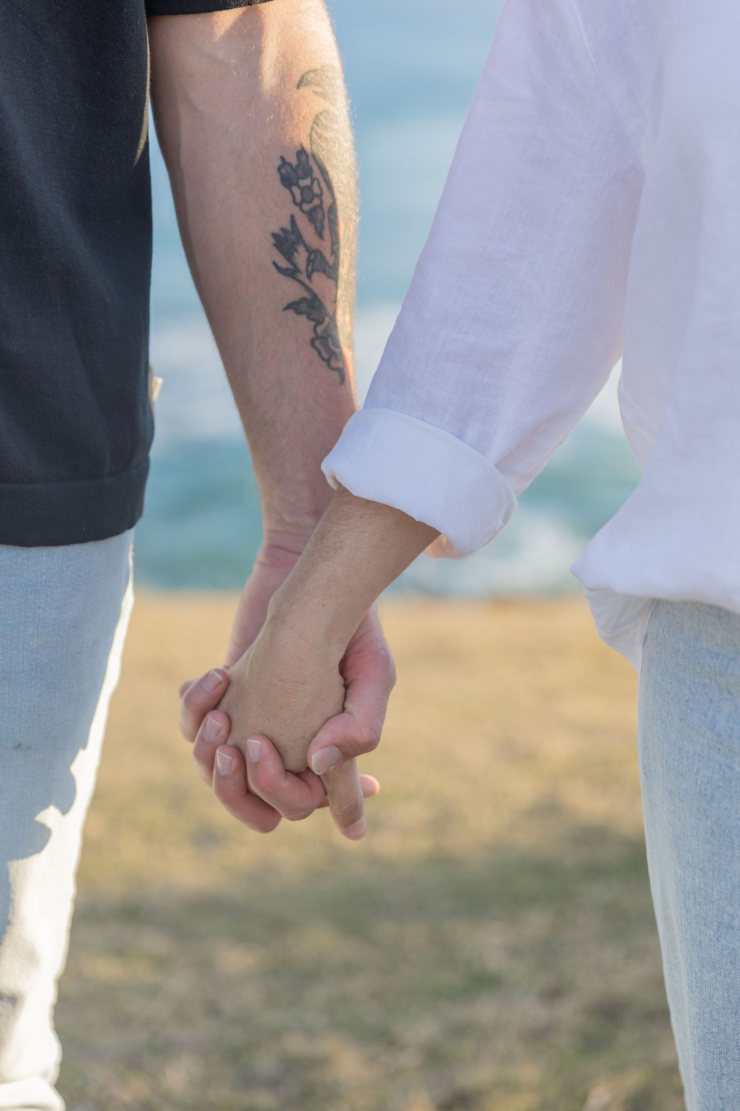 Close-up view of couple holding hands with floral tattoo visible on one person's forearm against a sunny outdoor background.
