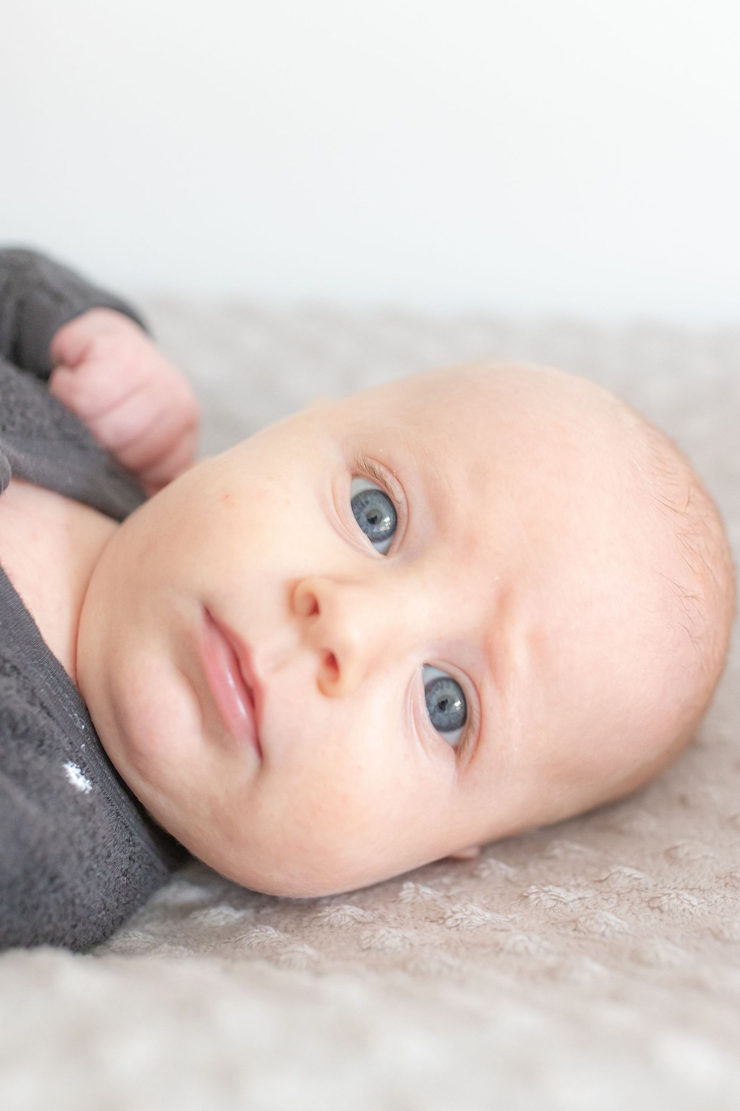 Adorable baby with blue eyes lying on a soft gray blanket, wearing a gray outfit, looking up curiously.