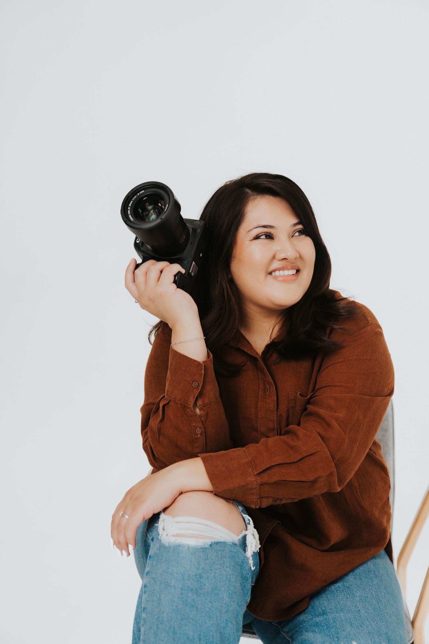 A photographer in a brown sweater and ripped jeans holds a professional camera while posing against a white backdrop.