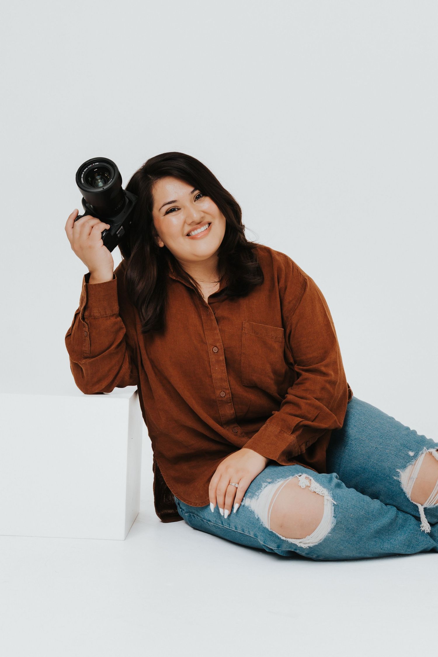 Photographer in brown button-down shirt and distressed jeans sits casually while holding camera against white background.