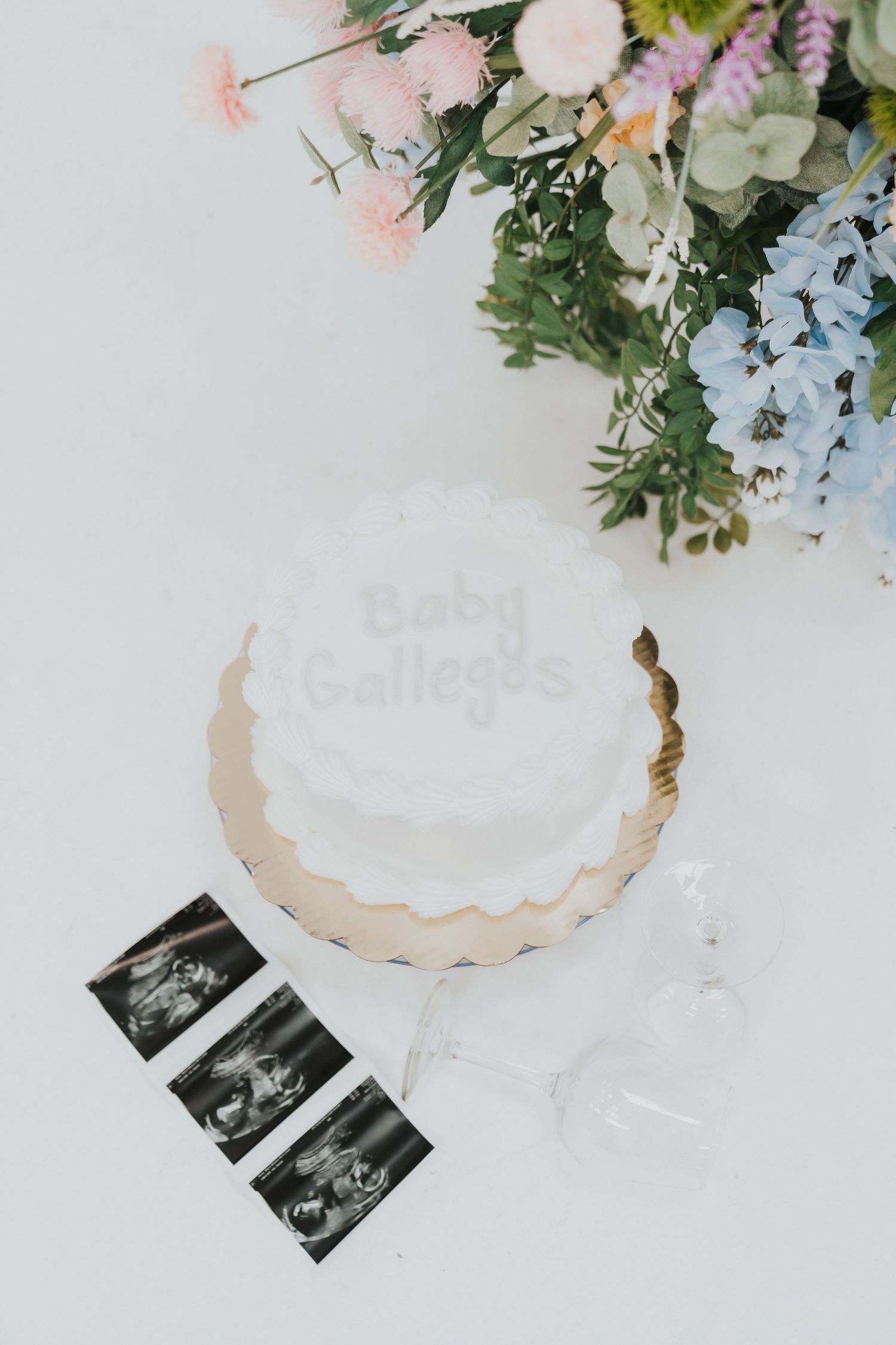 White cake with 'Happy Birthday' text alongside floral arrangement and ultrasound photos on white surface.