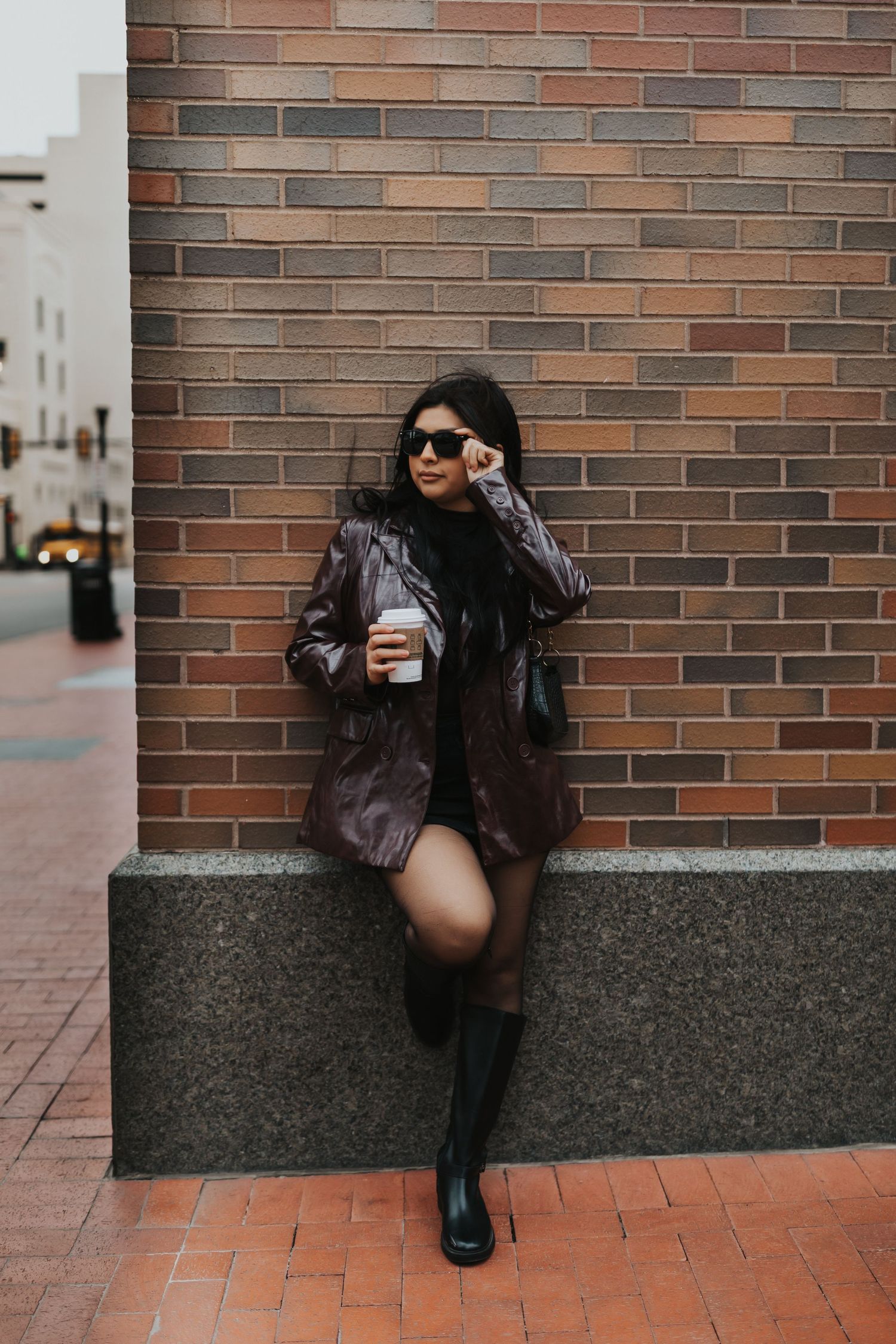 Stylish person in leather jacket and boots holding coffee while sitting on concrete ledge against brick wall.