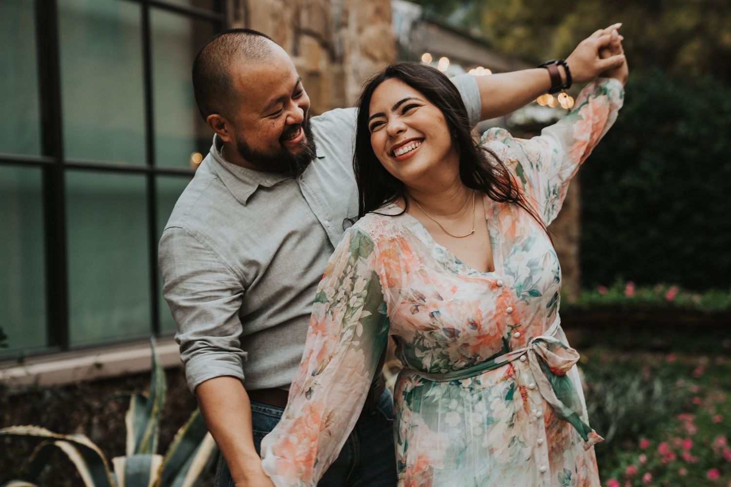 Joyful couple dancing together outdoors near a glass building, sharing an intimate candid moment in a garden setting.