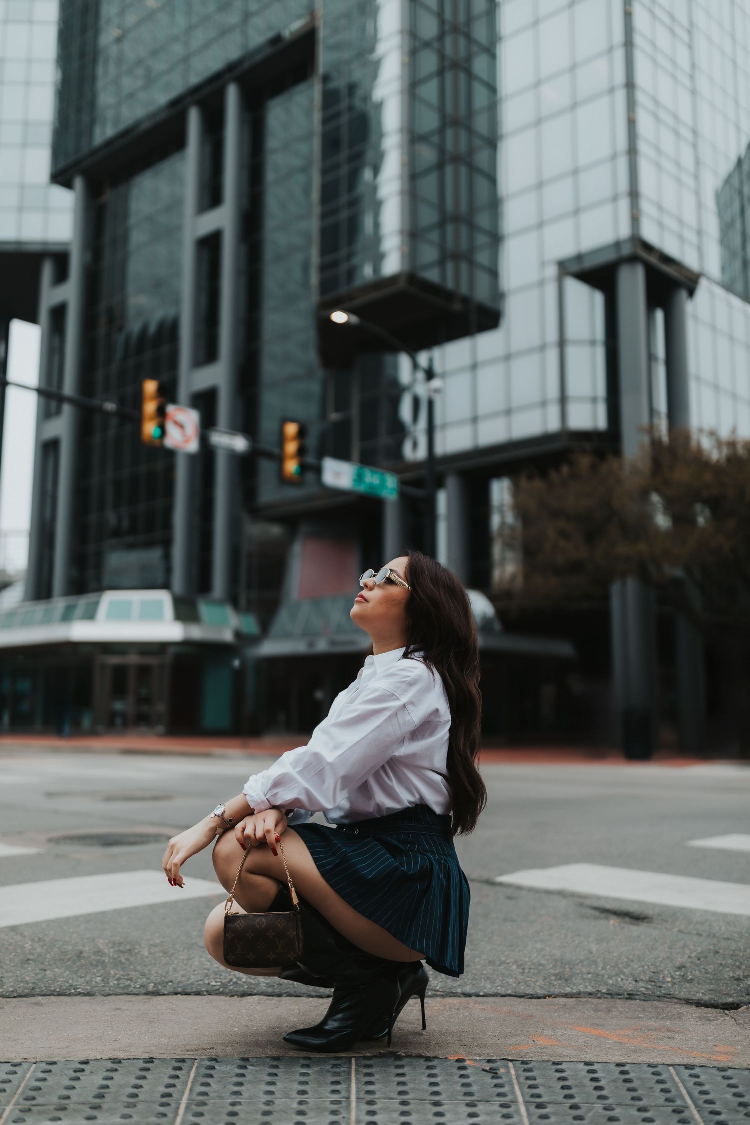 A person in a white shirt and skirt crouches on a city street with modern glass buildings in the background.