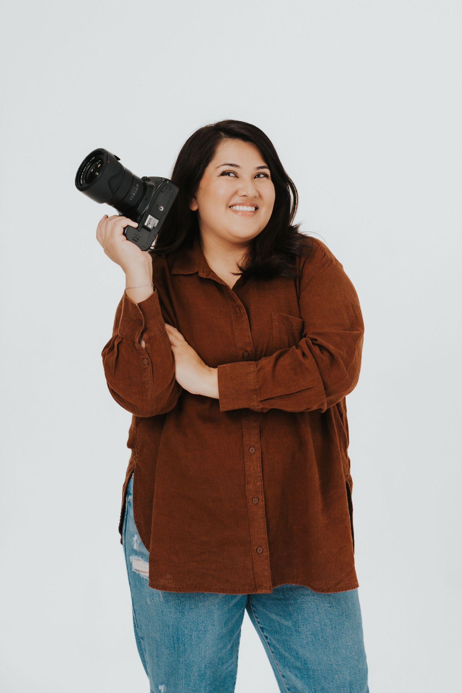Professional photographer wearing brown corduroy shirt and jeans holds DSLR camera against white background.