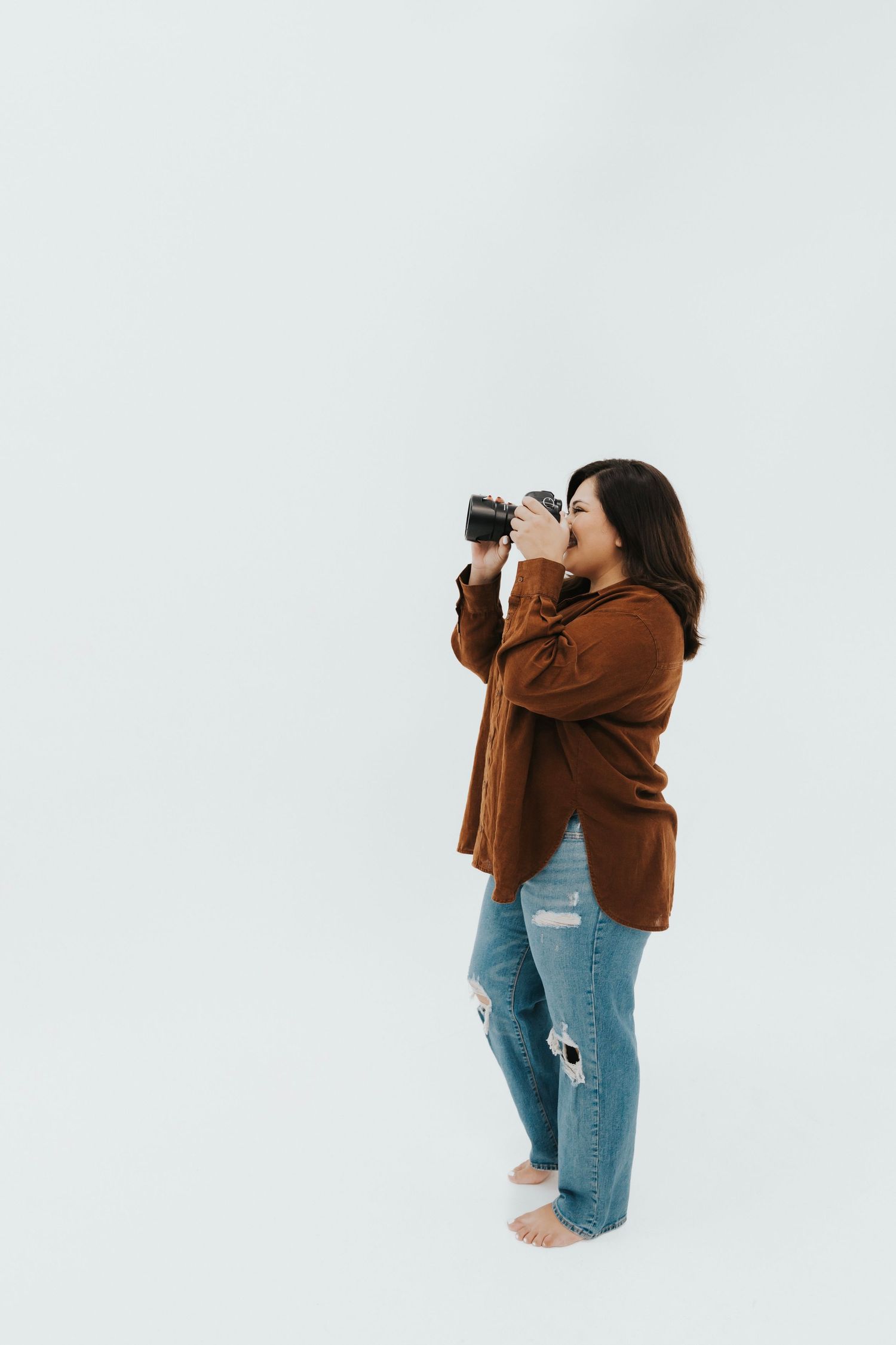 A photographer in a brown coat and ripped jeans takes pictures against a white background.