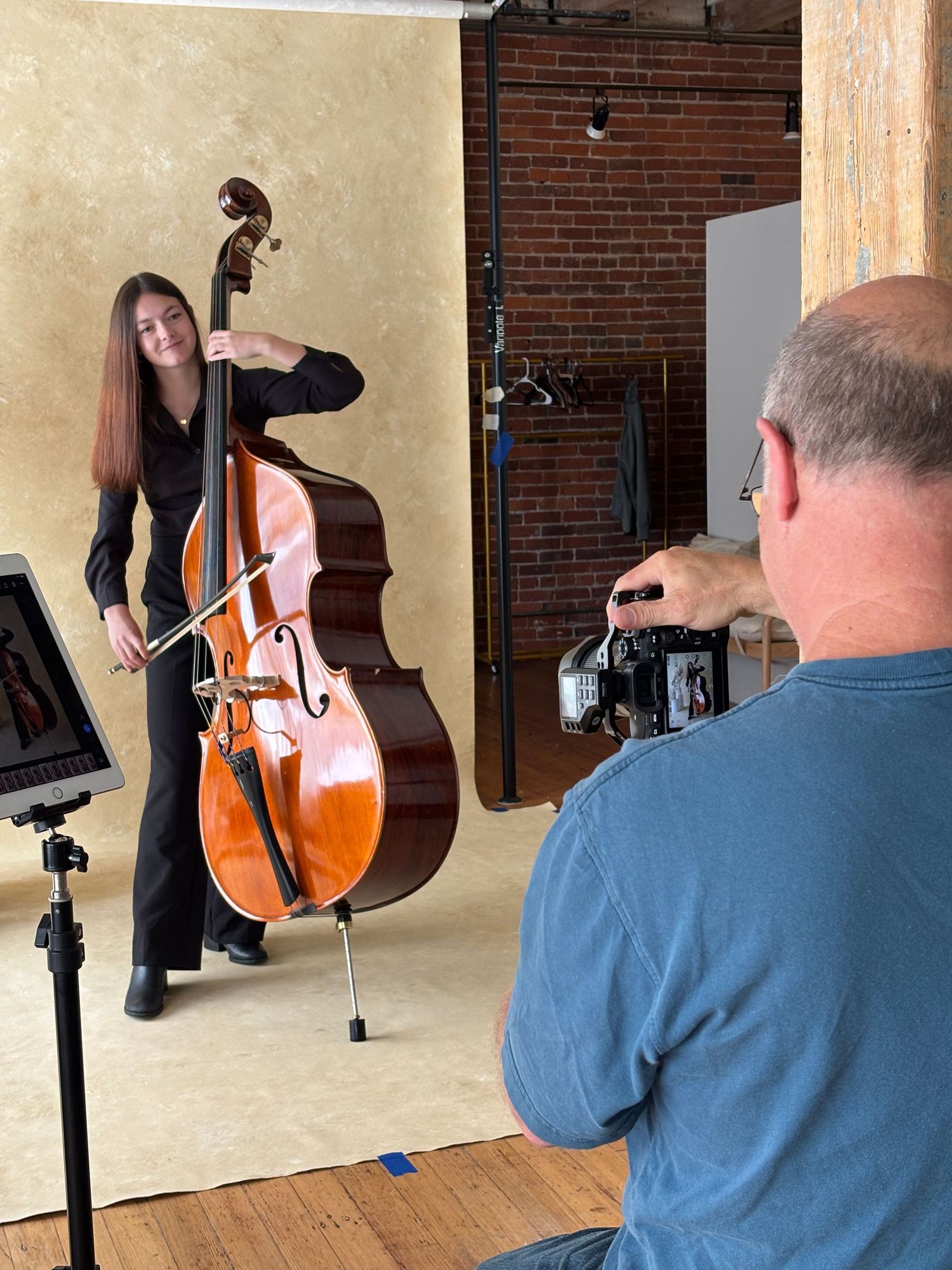 Musician's upright bass being photographed against a beige backdrop in a brick-walled studio setting.
