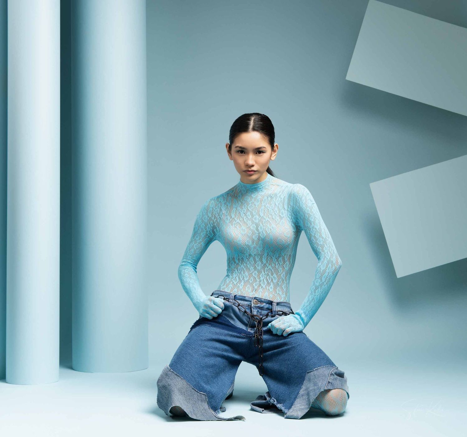 Fashion model wearing pastel blue sweater and ripped jeans kneels in minimalist studio setting.