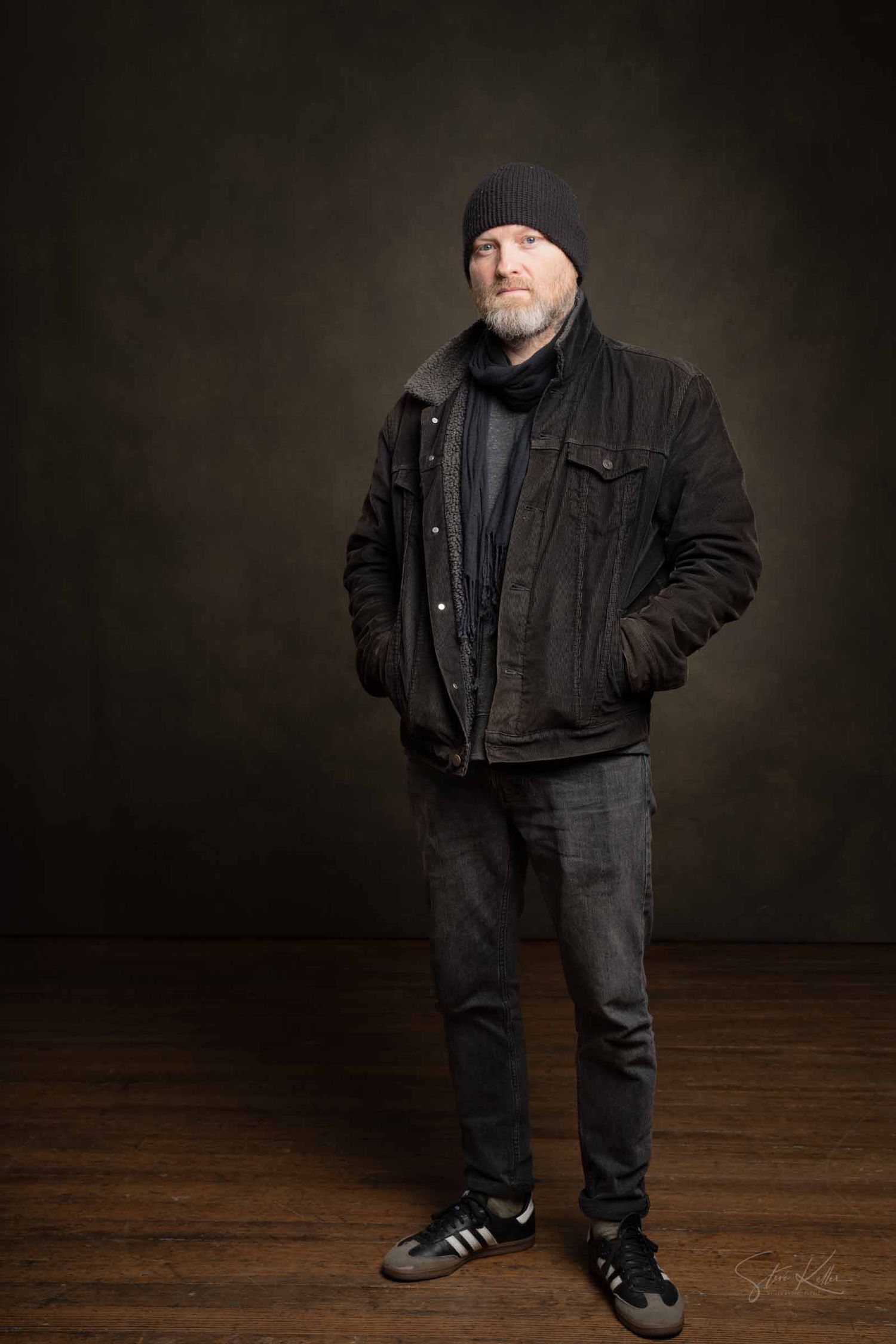 A figure wearing all black clothing stands against a dark backdrop on a wooden floor in a studio setting.
