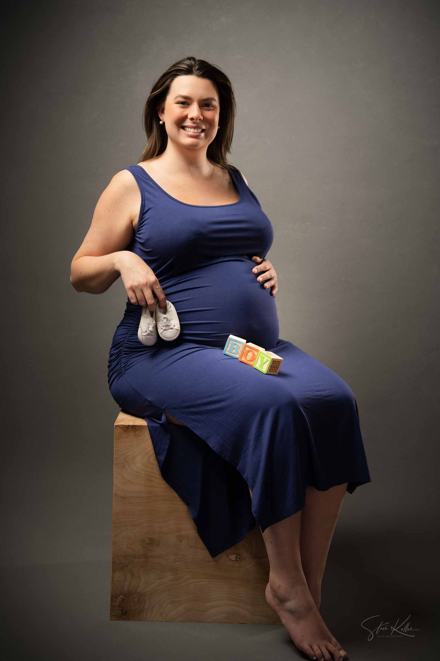 A pregnant woman in a navy dress poses on a wooden block for artistic maternity photos.