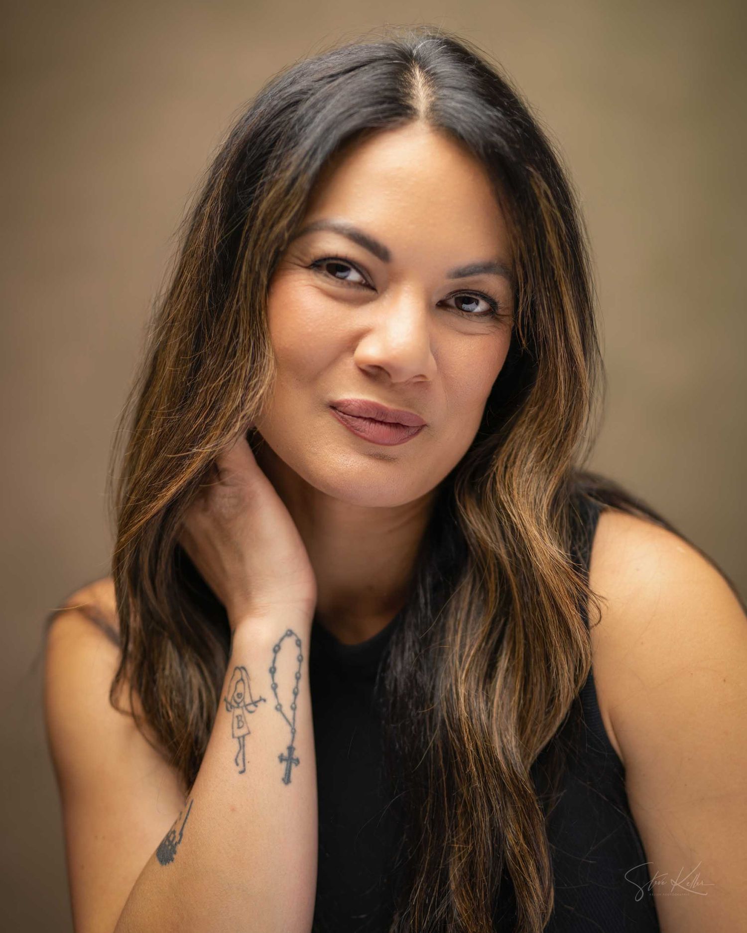 Woman with long dark hair and wrist tattoos poses for a portrait in a black sleeveless top.