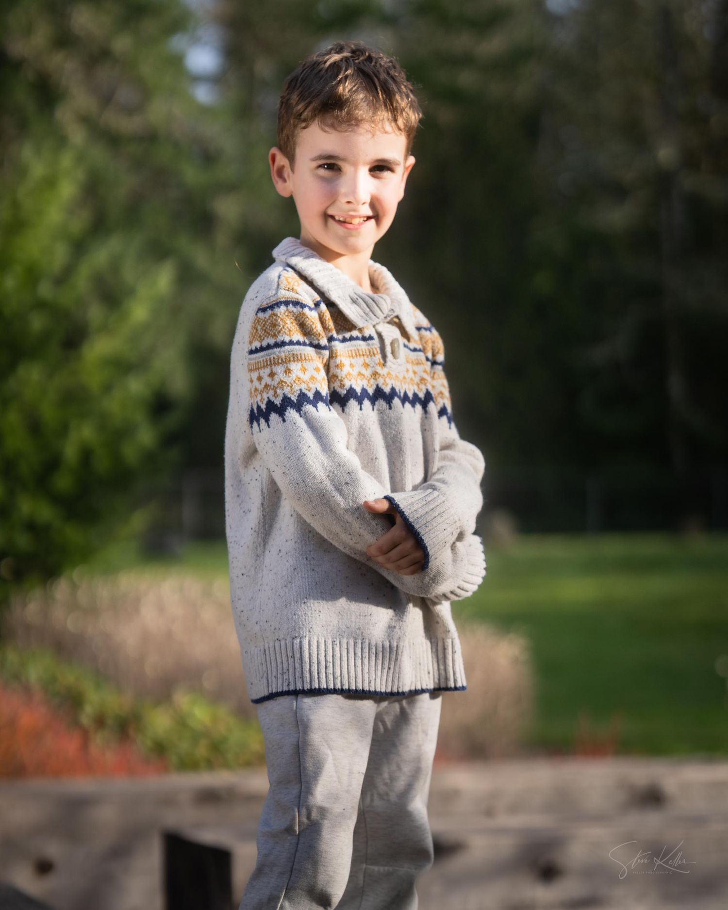 A child wearing a patterned knit sweater and gray pants stands outdoors against a blurred natural background.