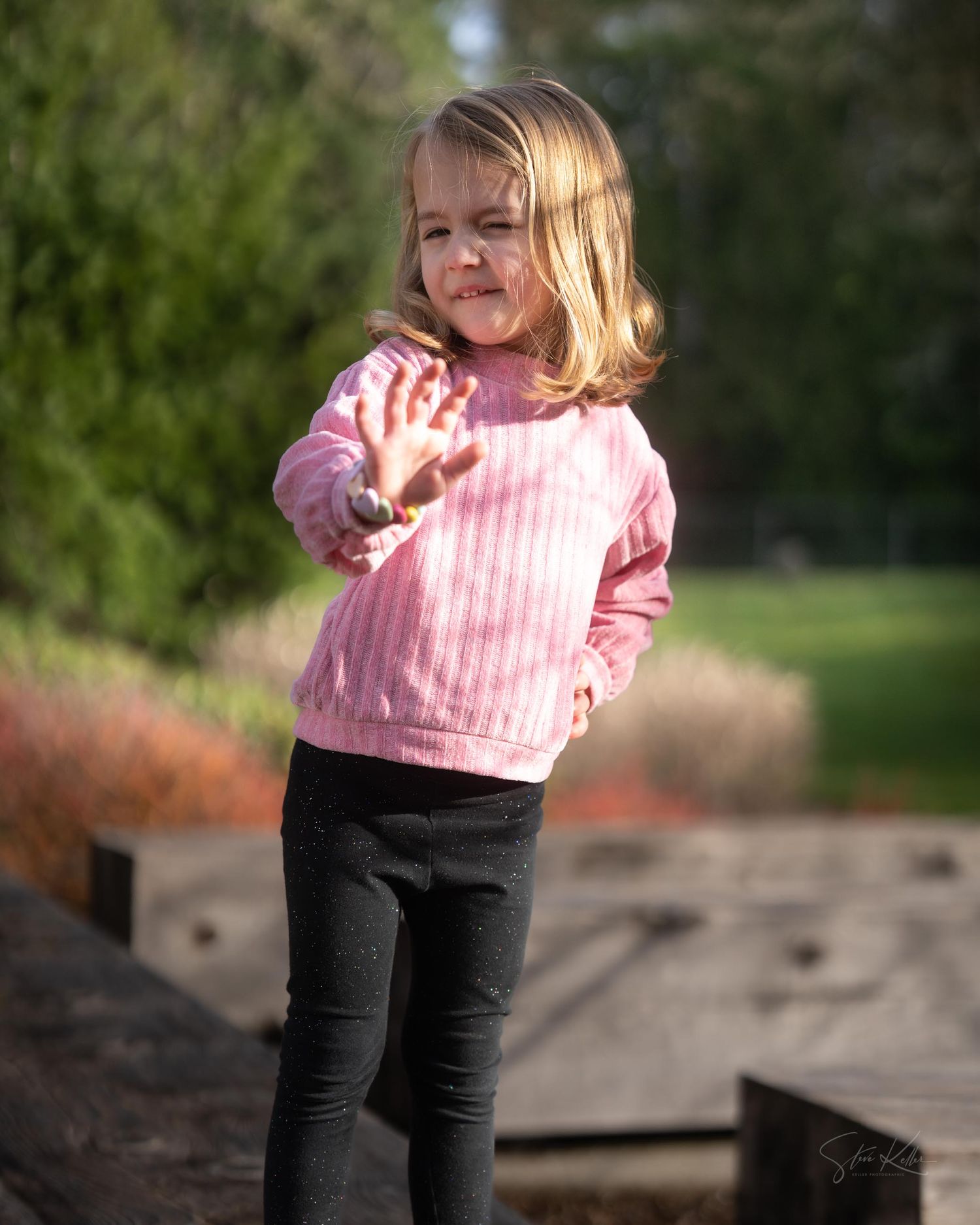 Young child in pink top and dark pants smiles while playing outside in a natural setting.