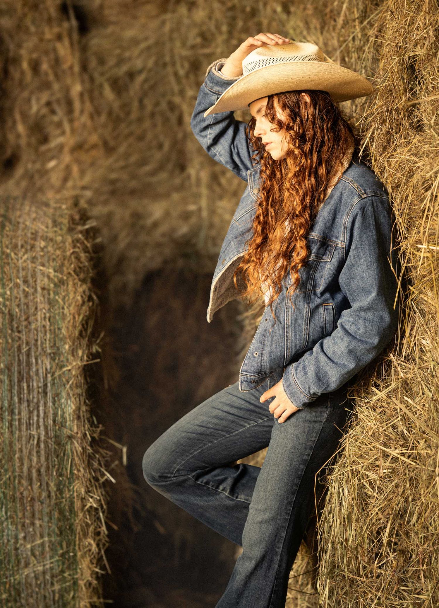 A young woman with curly red hair wearing a denim jacket and cowboy hat leans against hay bales outdoors.