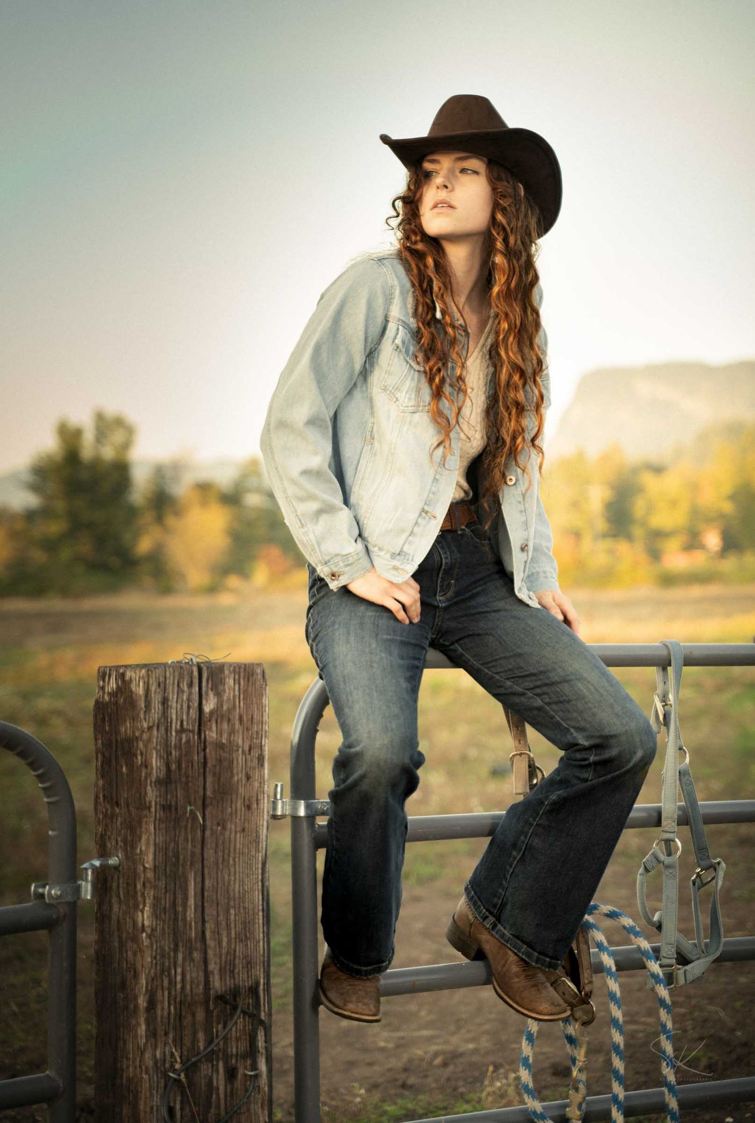Young woman in cowboy hat, denim jacket, and jeans sitting on a ranch fence with curly hair and boots.