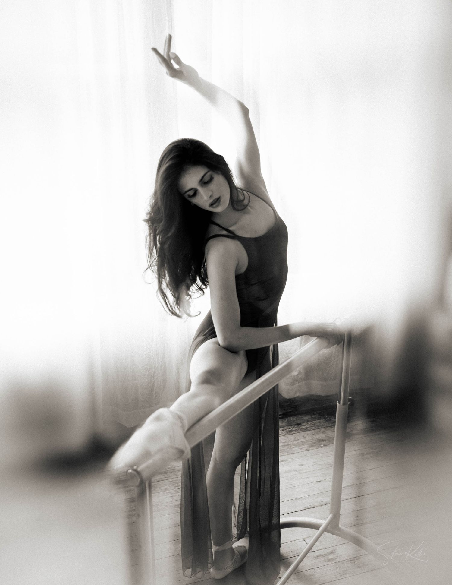 Graceful ballerina poses at ballet barre in black dance attire and white tutu in dramatic black and white photograph.