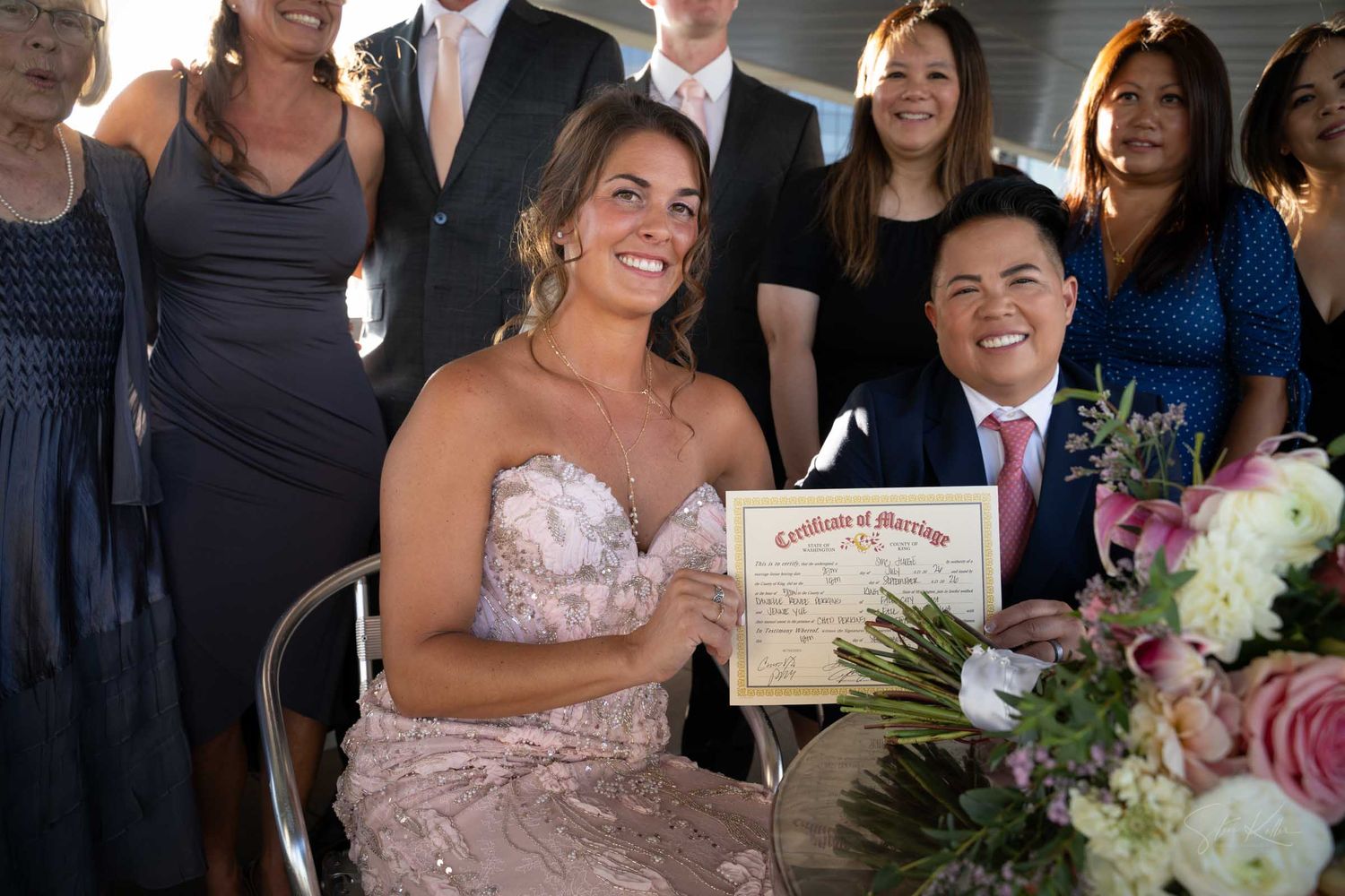 Newlyweds smile holding marriage certificate surrounded by guests at wedding reception.