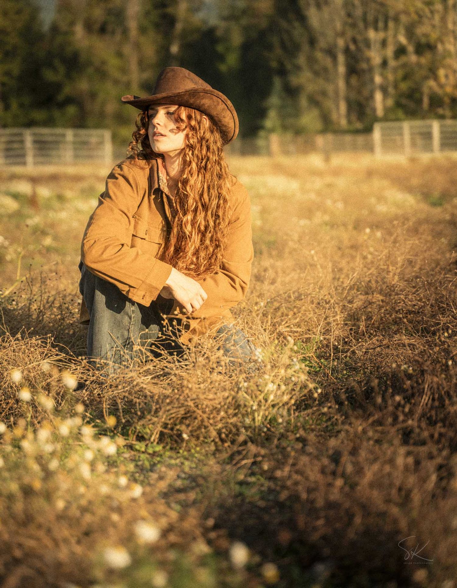 A woman with long curly red hair wearing a brown cowboy hat and jacket sits in a golden field at sunset.