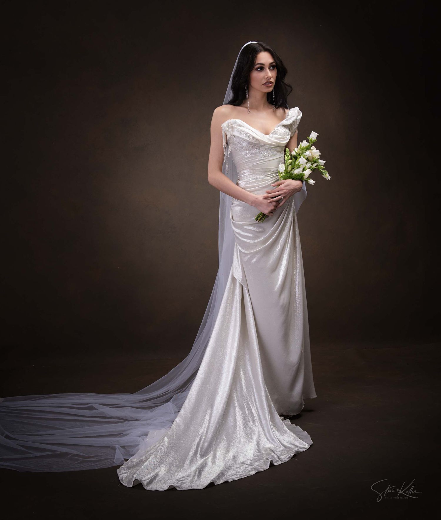 Bride in flowing white dress poses gracefully with white lilies against moody dark background.