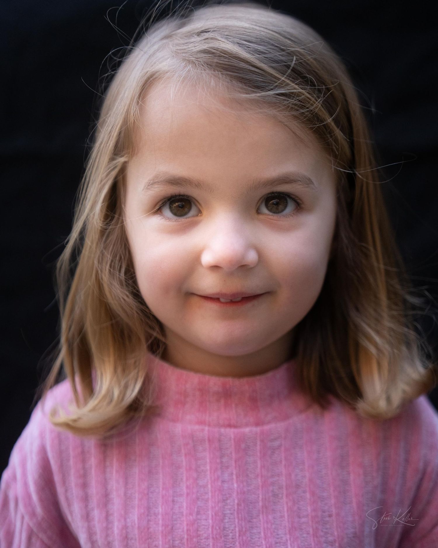 Young child in a pink sweater smiling warmly at the camera against a dark background.