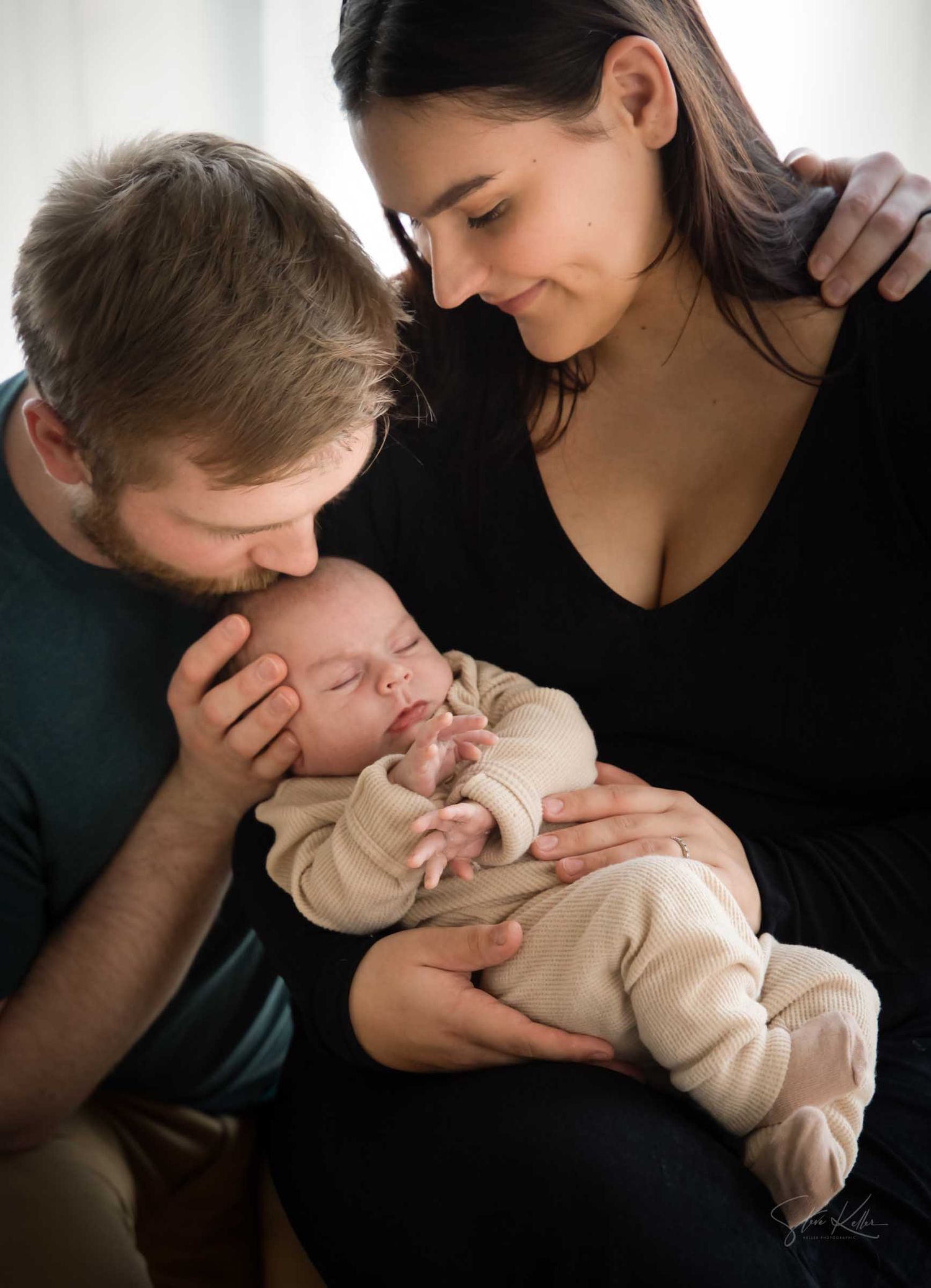 New parents tenderly kissing and gazing at their sleeping newborn baby dressed in cream outfit.