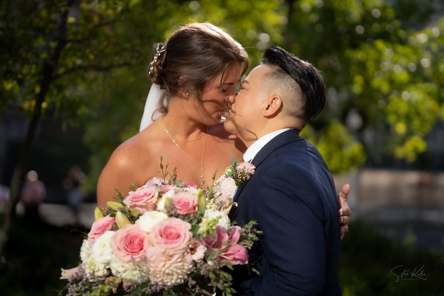 Two brides share a tender moment, one in a white gown holding pink roses, the other in a navy suit, surrounded by lush greenery.