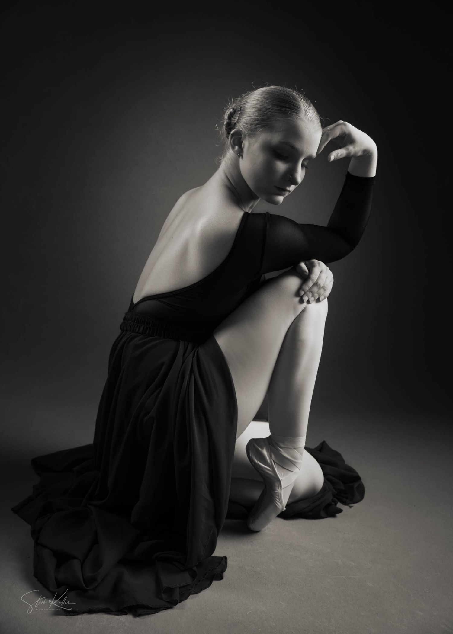 A ballet dancer in black dress poses elegantly on the floor in dramatic lighting.