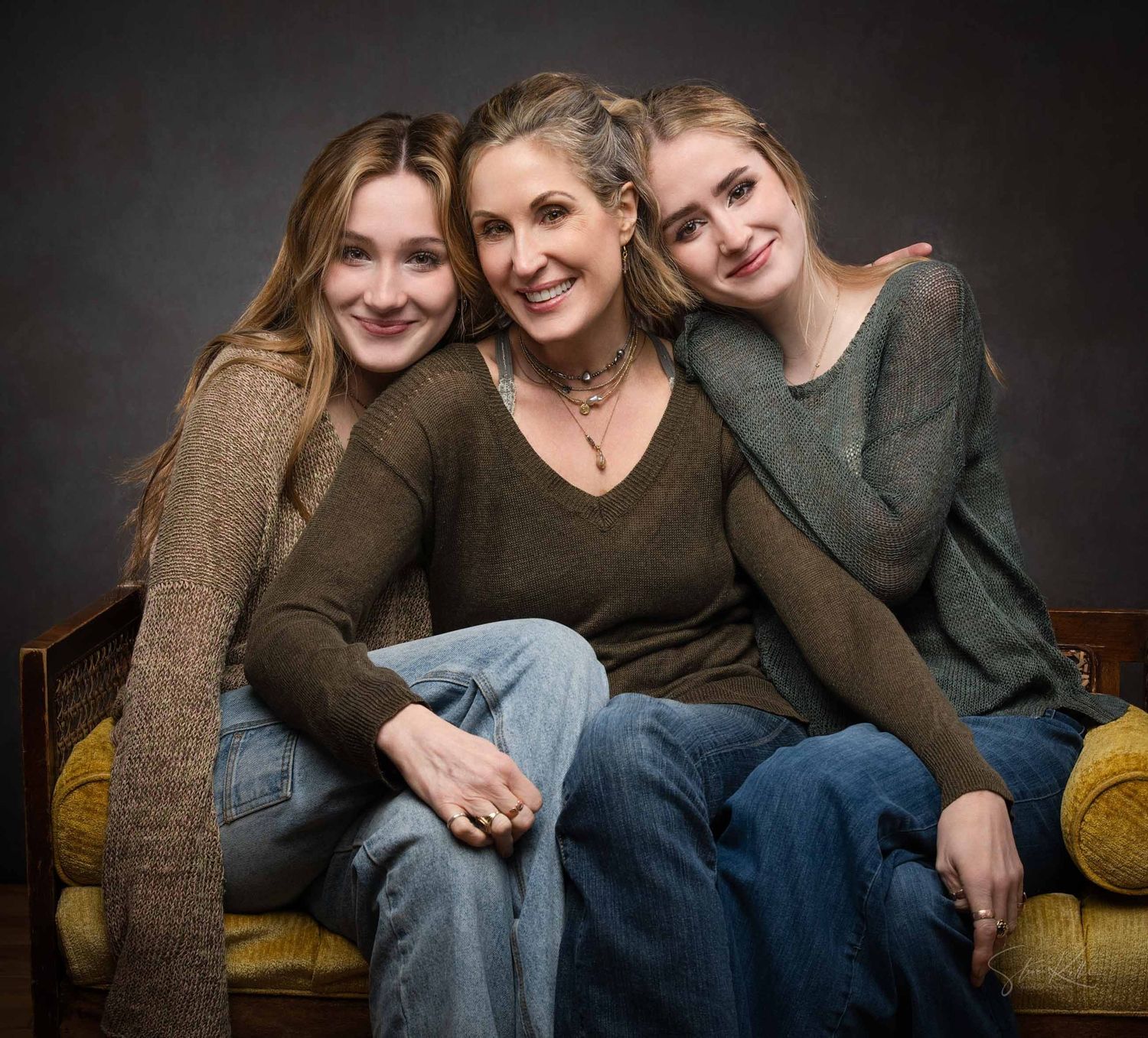 Three friends in casual sweaters pose together on a brown couch against a dark background in a studio setting.