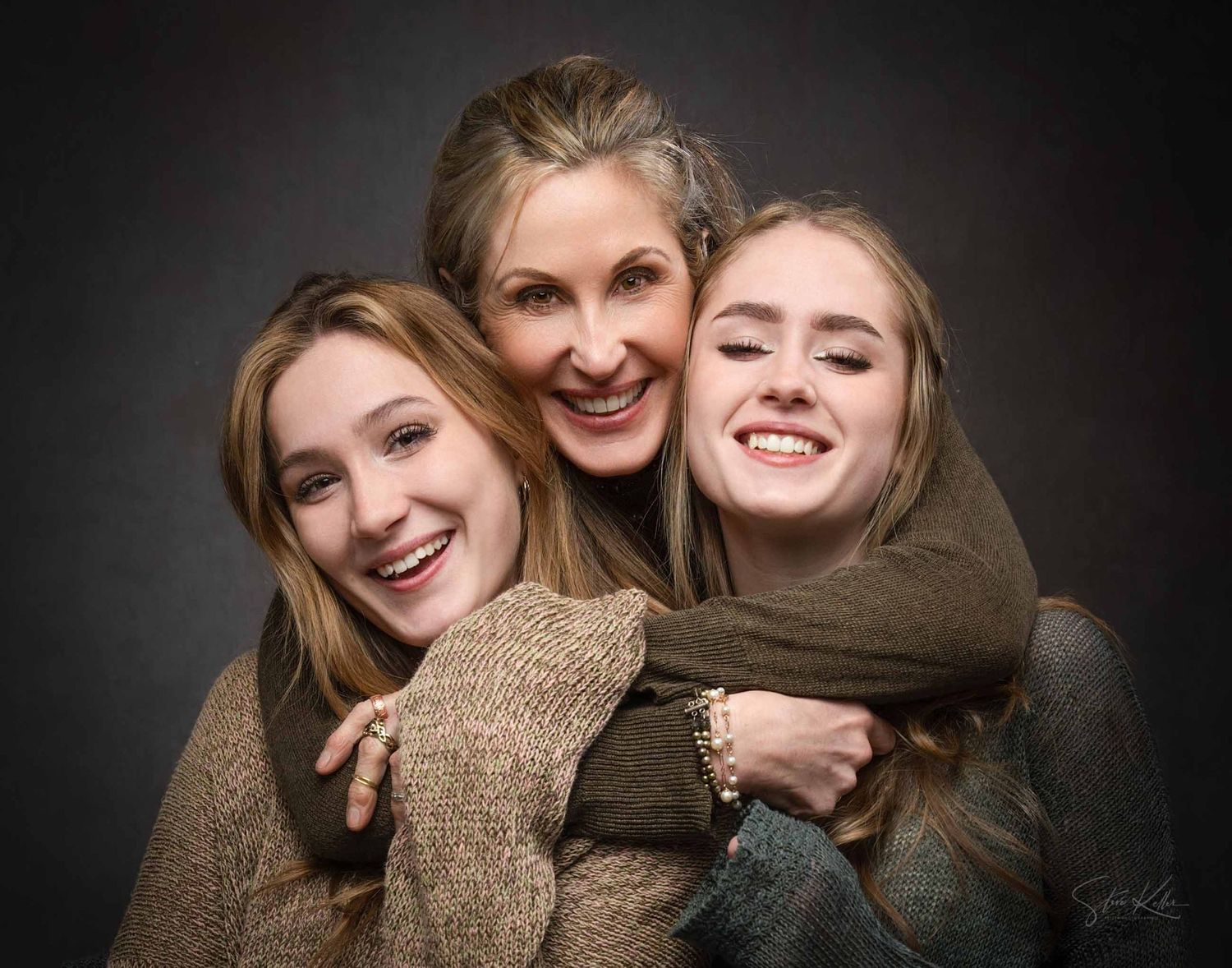 Three people wearing brown sweaters embrace and smile warmly in a professional portrait against a dark background.