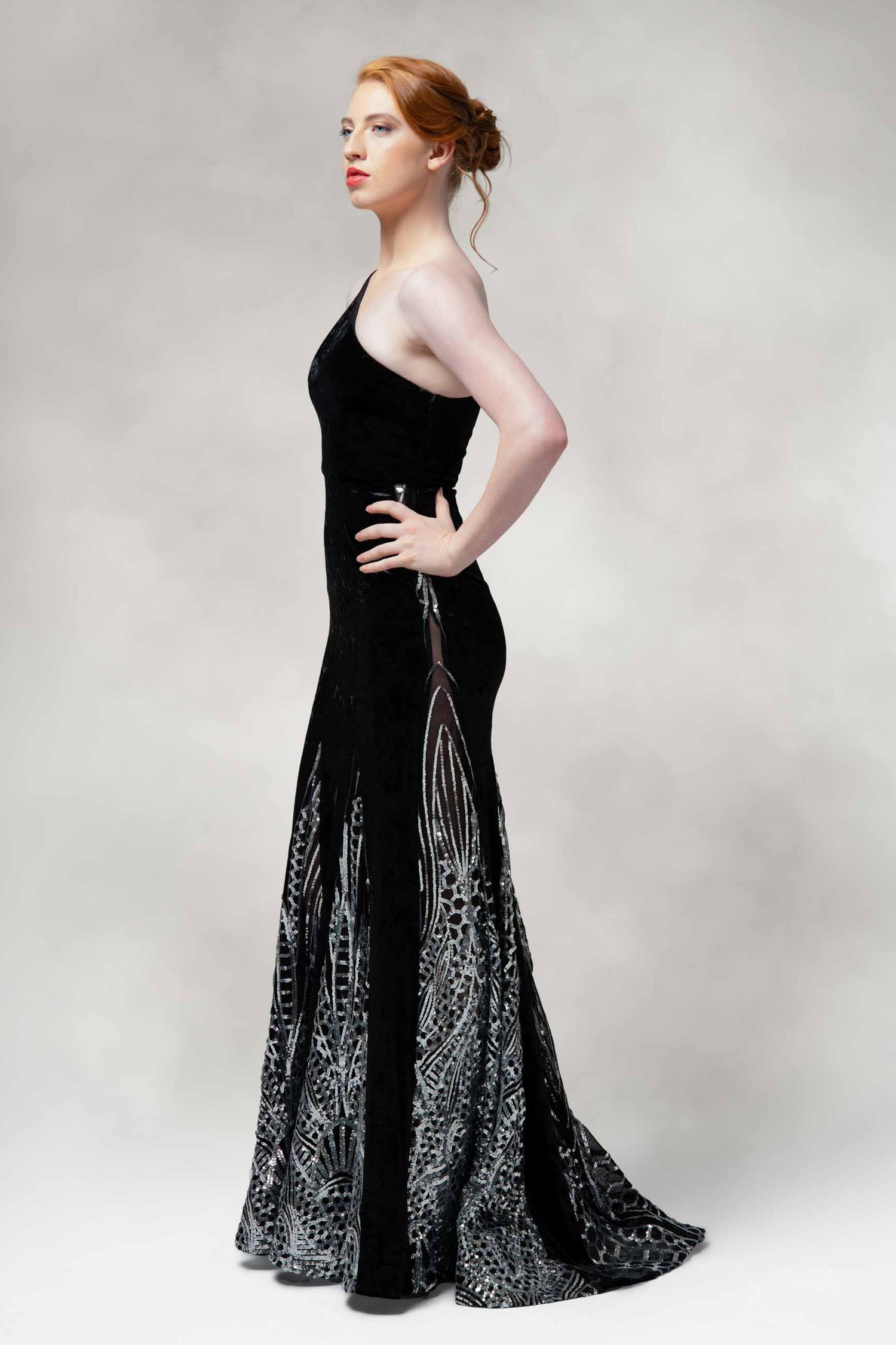 Fashion model wearing a black evening gown with silver sequined details against grey background.