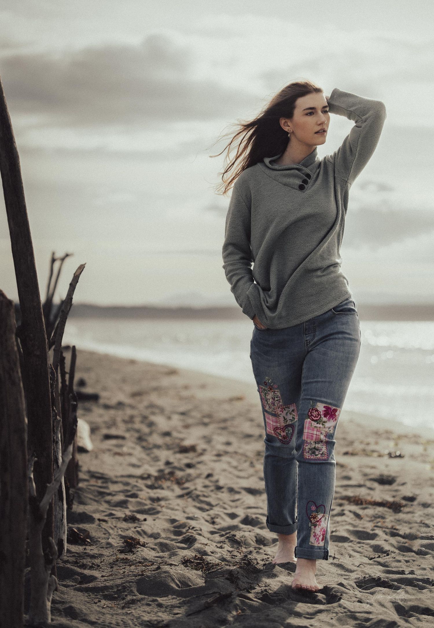 Person in a gray sweater and patched jeans stands thoughtfully on a misty beach with wind-blown hair at sunset.