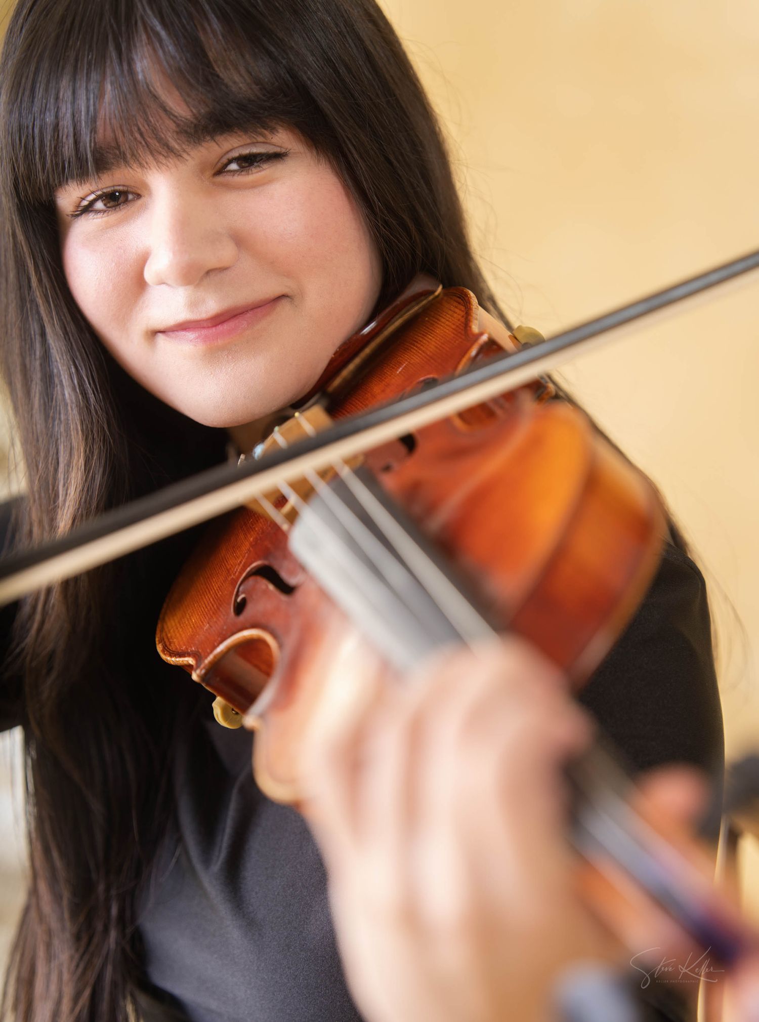 A violinist holds their wooden instrument and bow while smiling warmly against a neutral background.