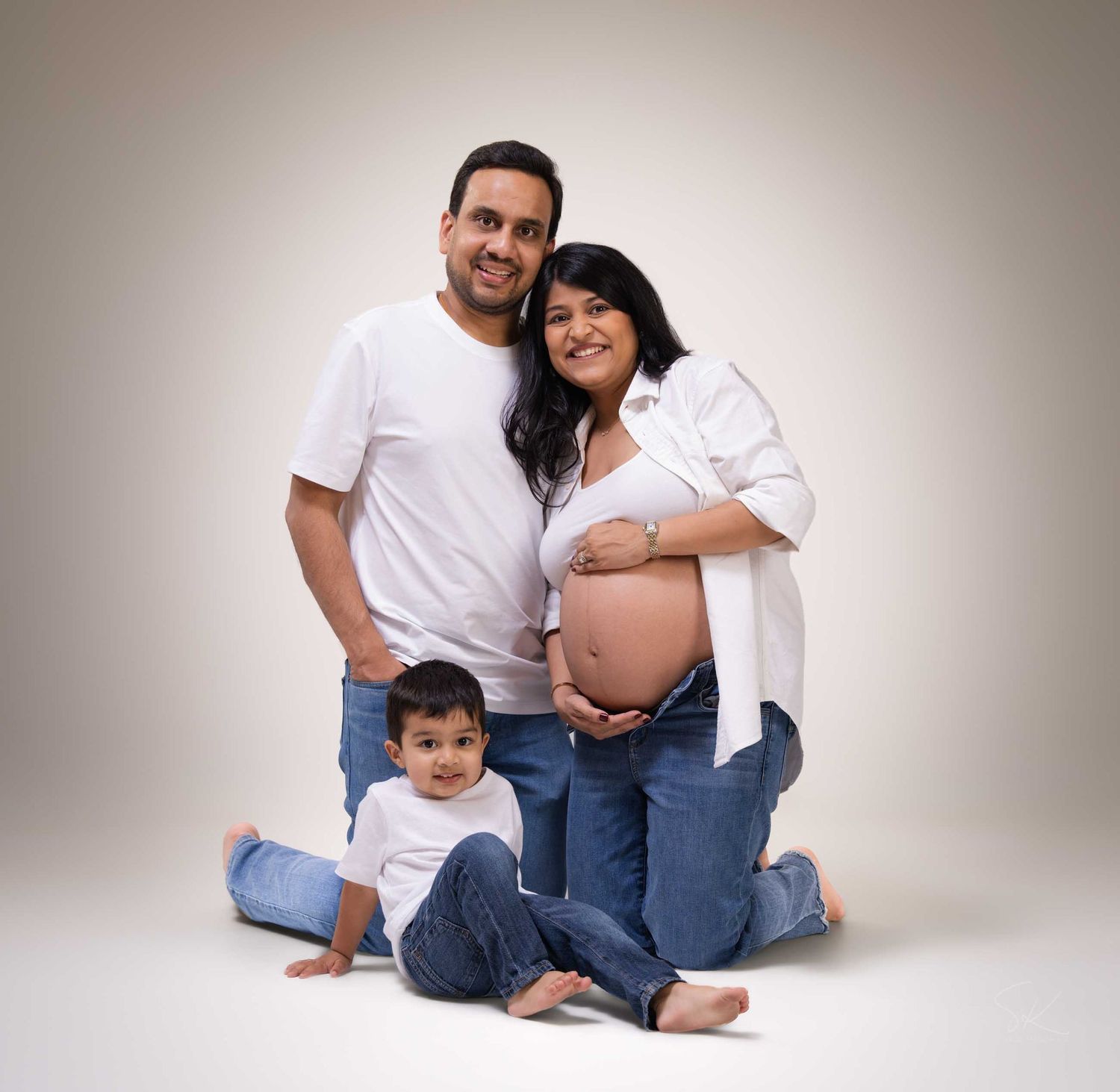 Pregnant woman in white shirt with husband and toddler son posing for maternity family portrait in studio.