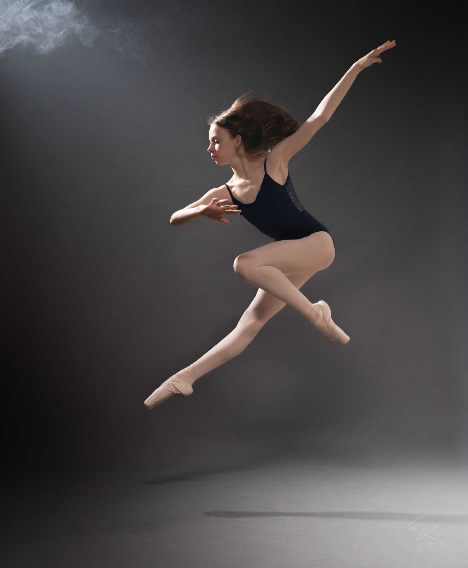 A ballet dancer leaps gracefully in mid-air wearing a black leotard and pointe shoes against a dark smoky background.