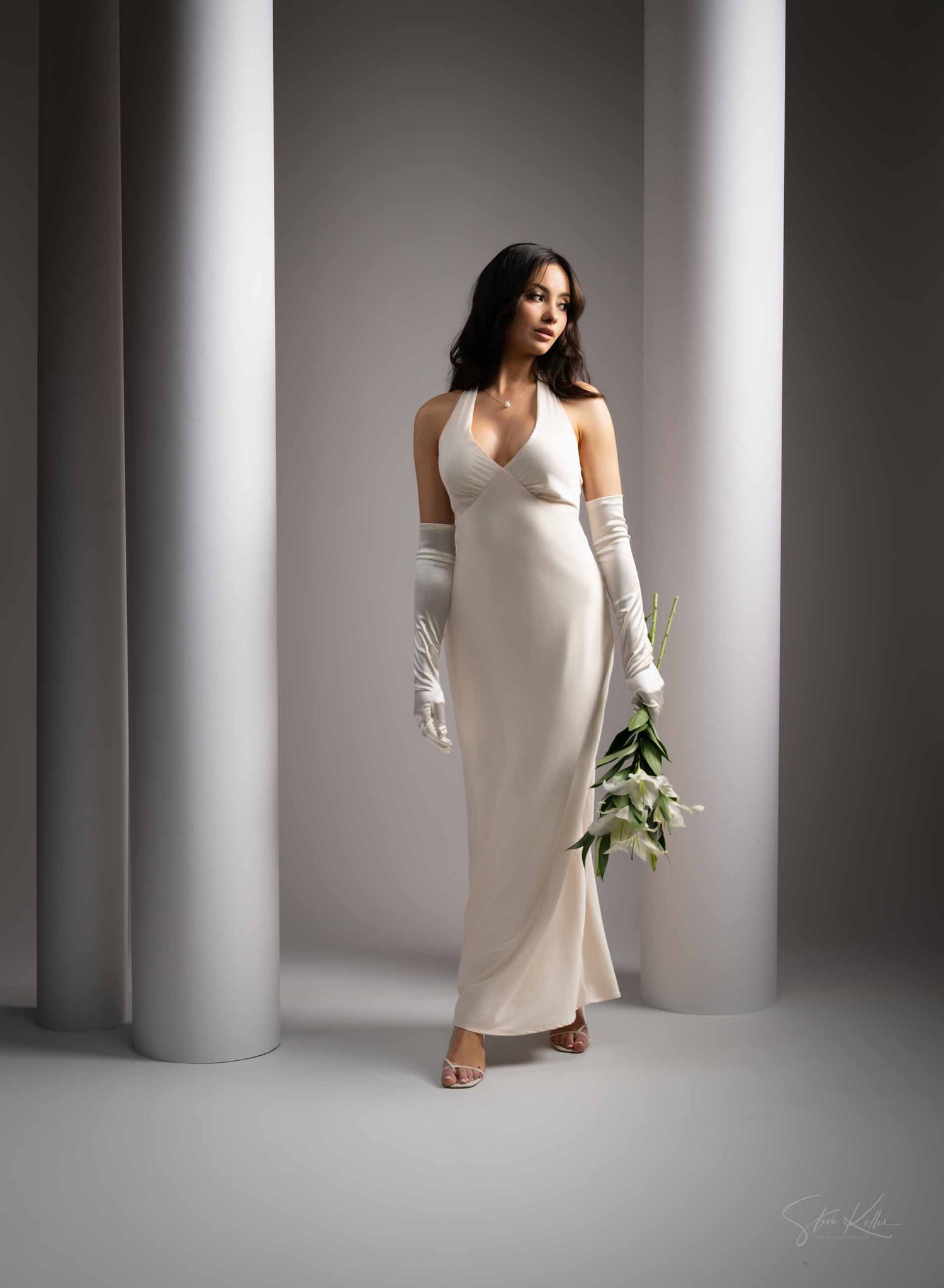 A woman in a sleek white dress and long gloves poses elegantly between modern white columns in a studio setting.