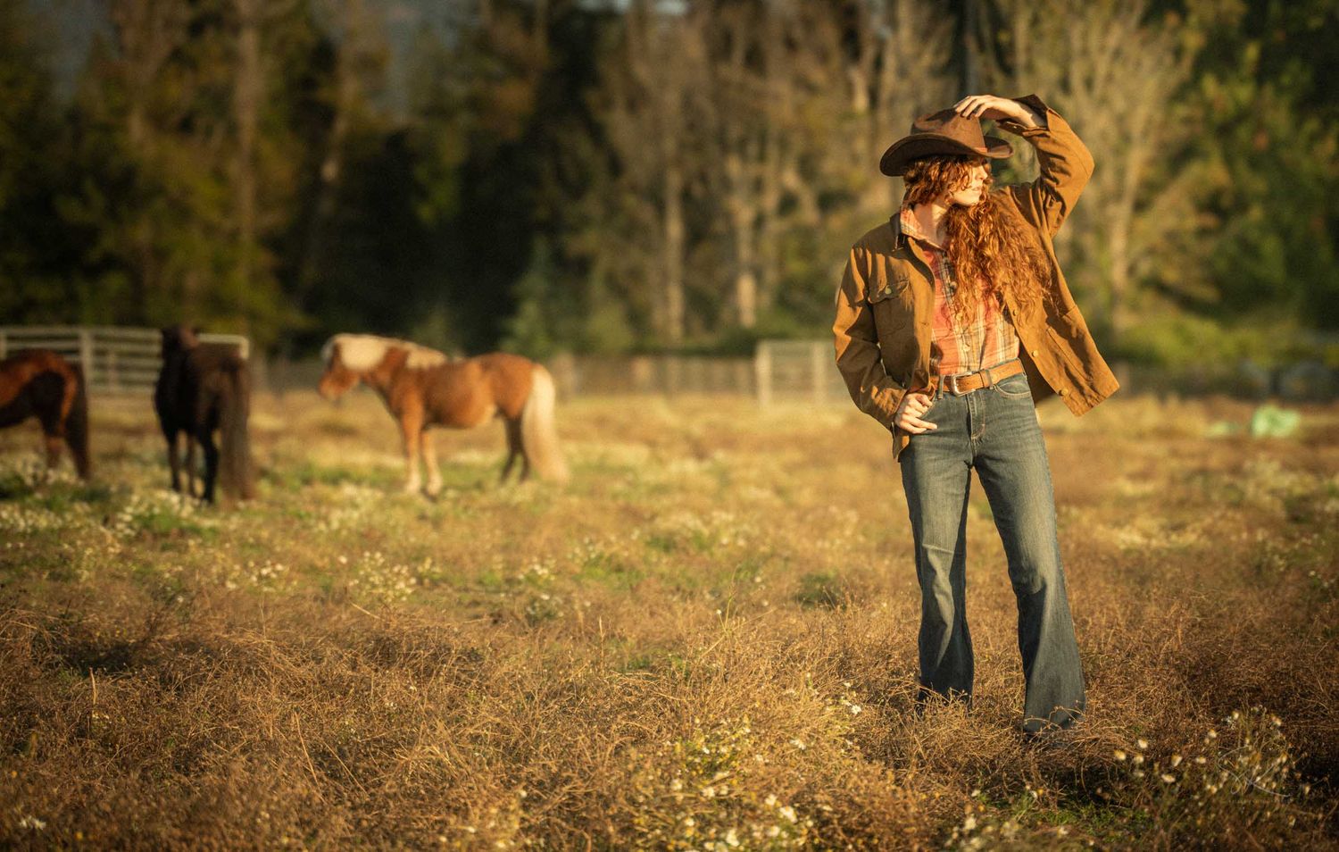 Woman in cowboy hat and flared jeans poses in golden field with horses grazing in background.