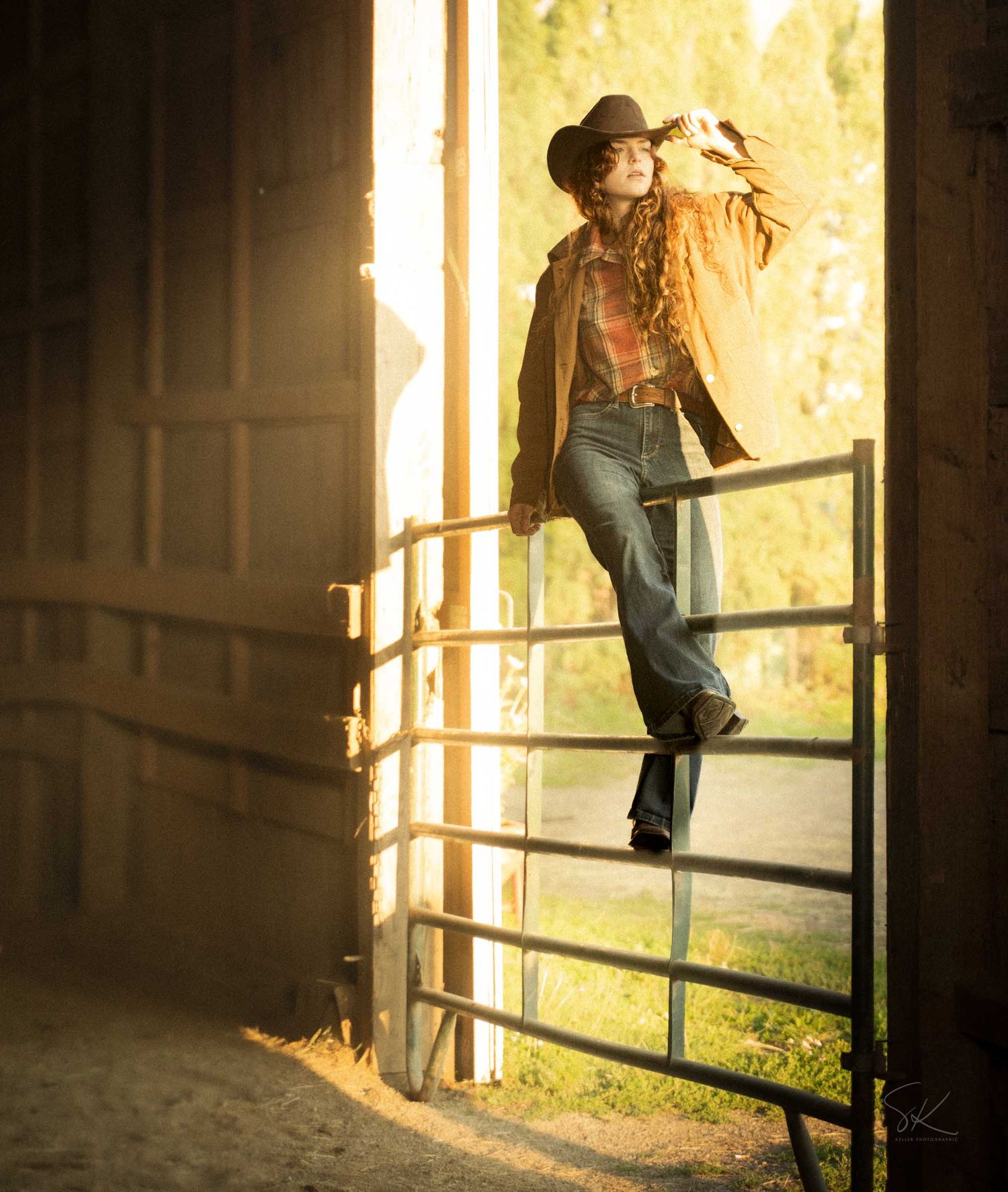 A woman in a cowboy hat and flare jeans poses on a metal gate inside a sunlit barn doorway.