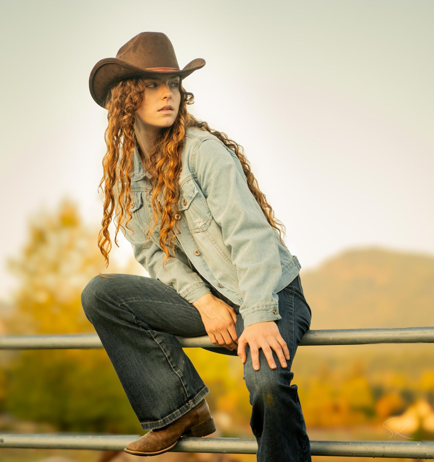 Young woman in cowboy hat, curly red hair, denim jacket, and jeans sitting on a fence in a rural outdoor setting.