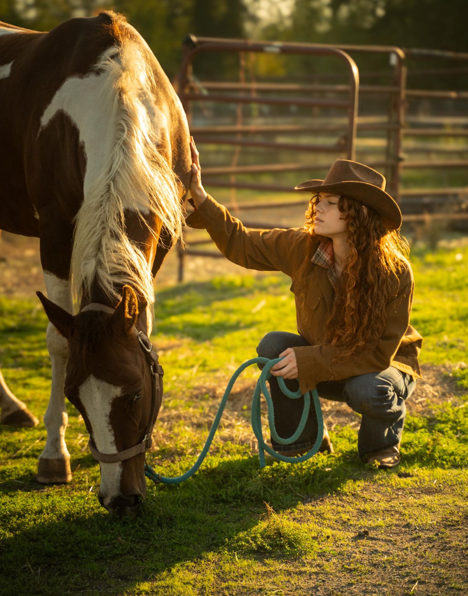 A cowgirl in a brown jacket and hat crouches beside a paint horse grazing in a sunlit green pasture.