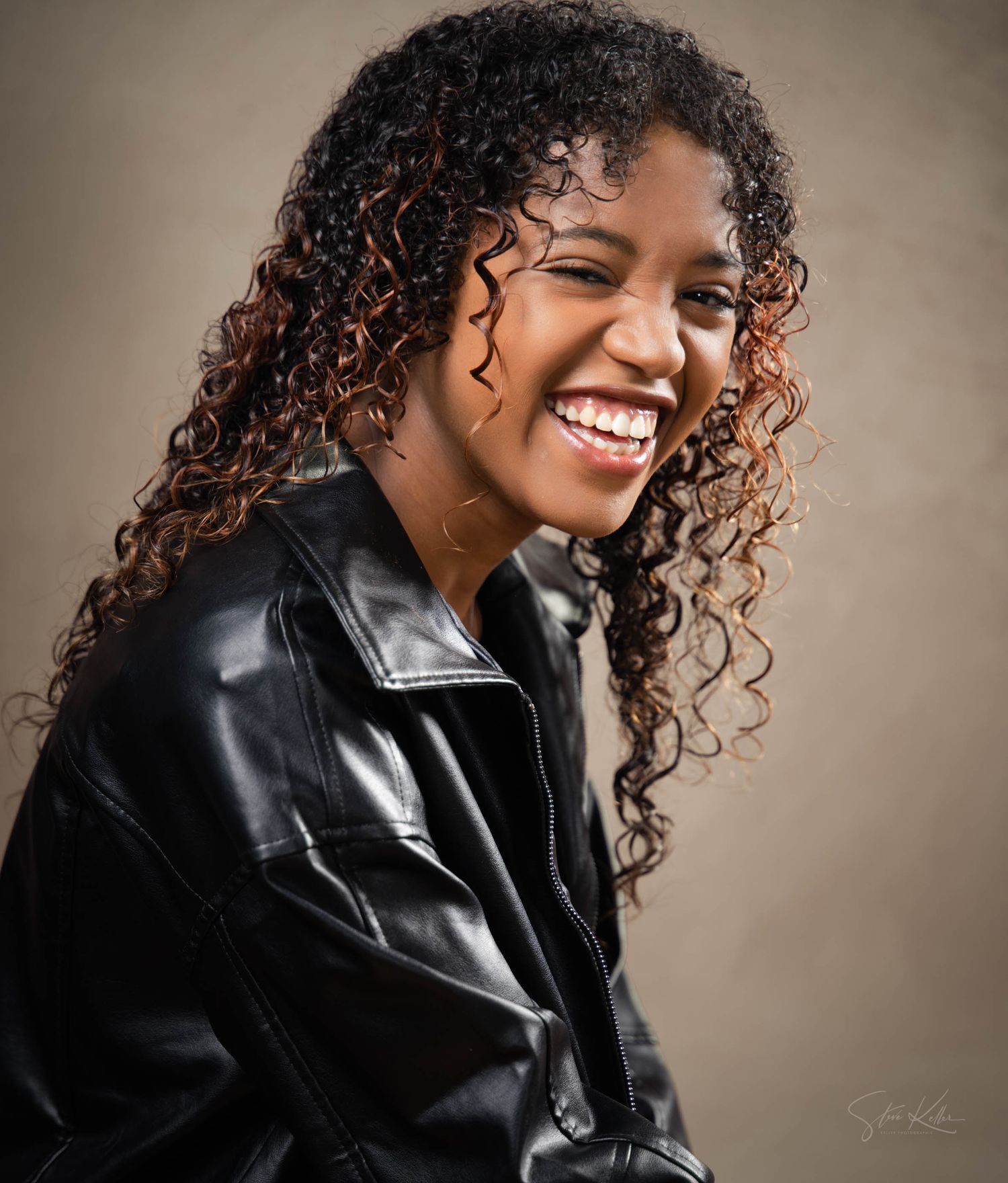 A person with curly hair wearing a black leather jacket smiles and poses against a neutral studio backdrop.