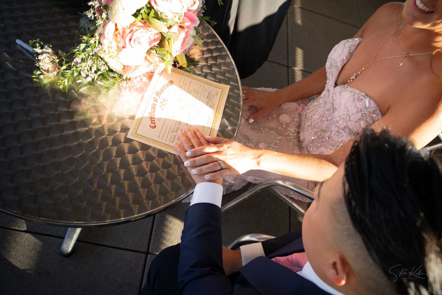 Aerial view of two brides holding hands at a glass table, one in a navy suit and the other in a pink lace gown, reading vows.